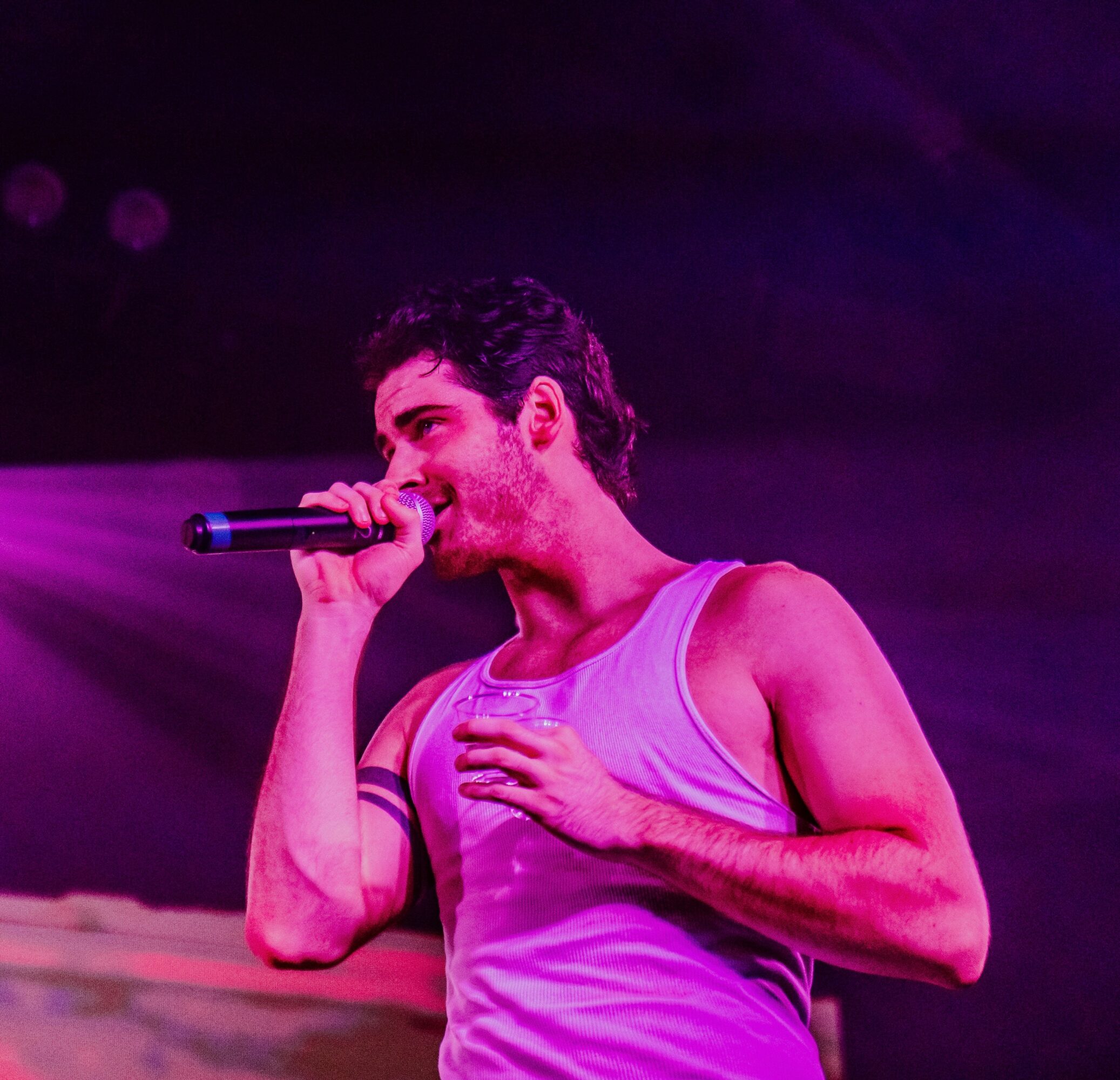 Person singing into a microphone on stage, wearing a sleeveless shirt, with hand on chest, under purple lighting.