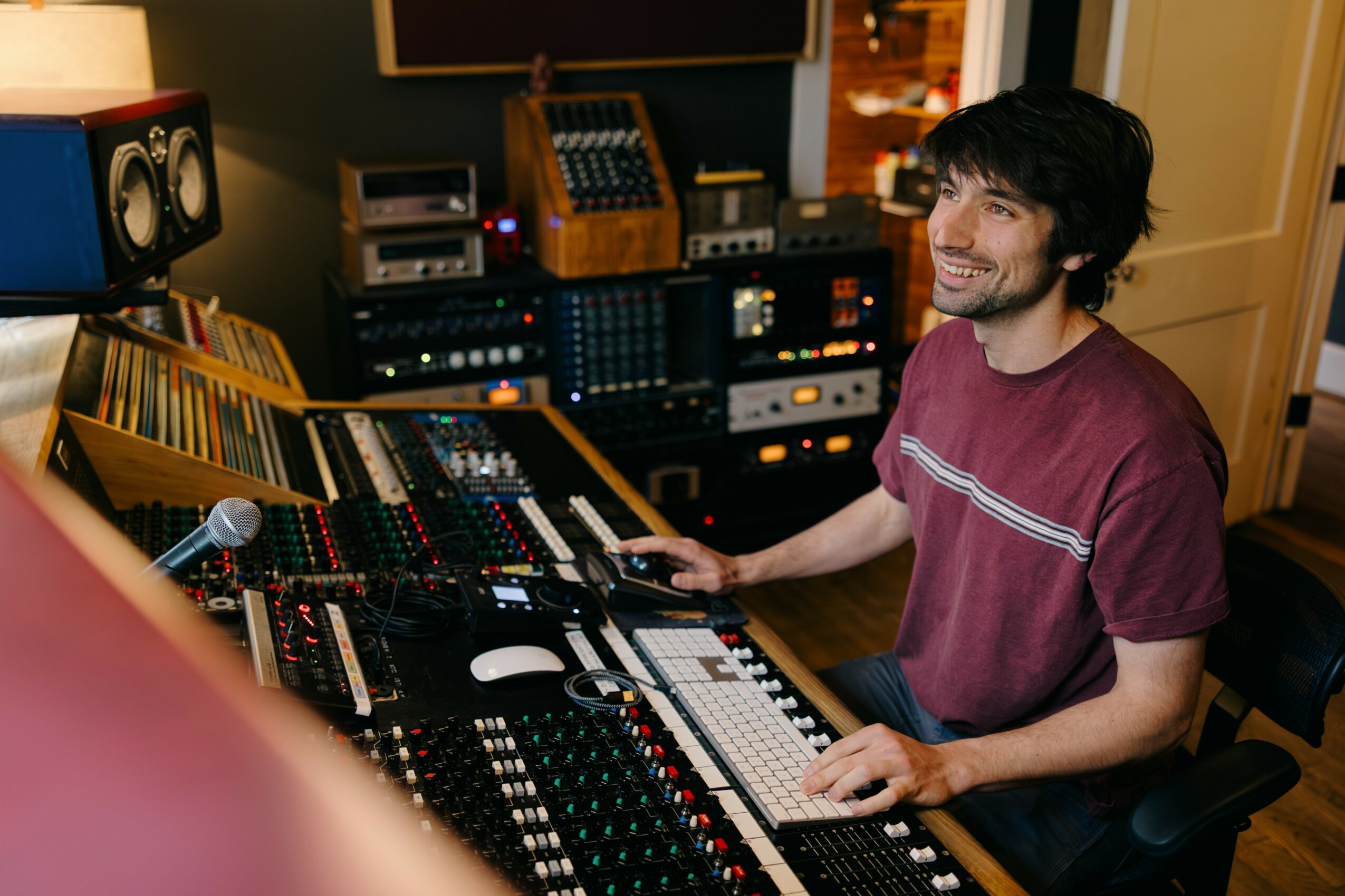 Young man with dark hair and beard in a recording studio, smiling, sitting at a mixing console with audio equipment.