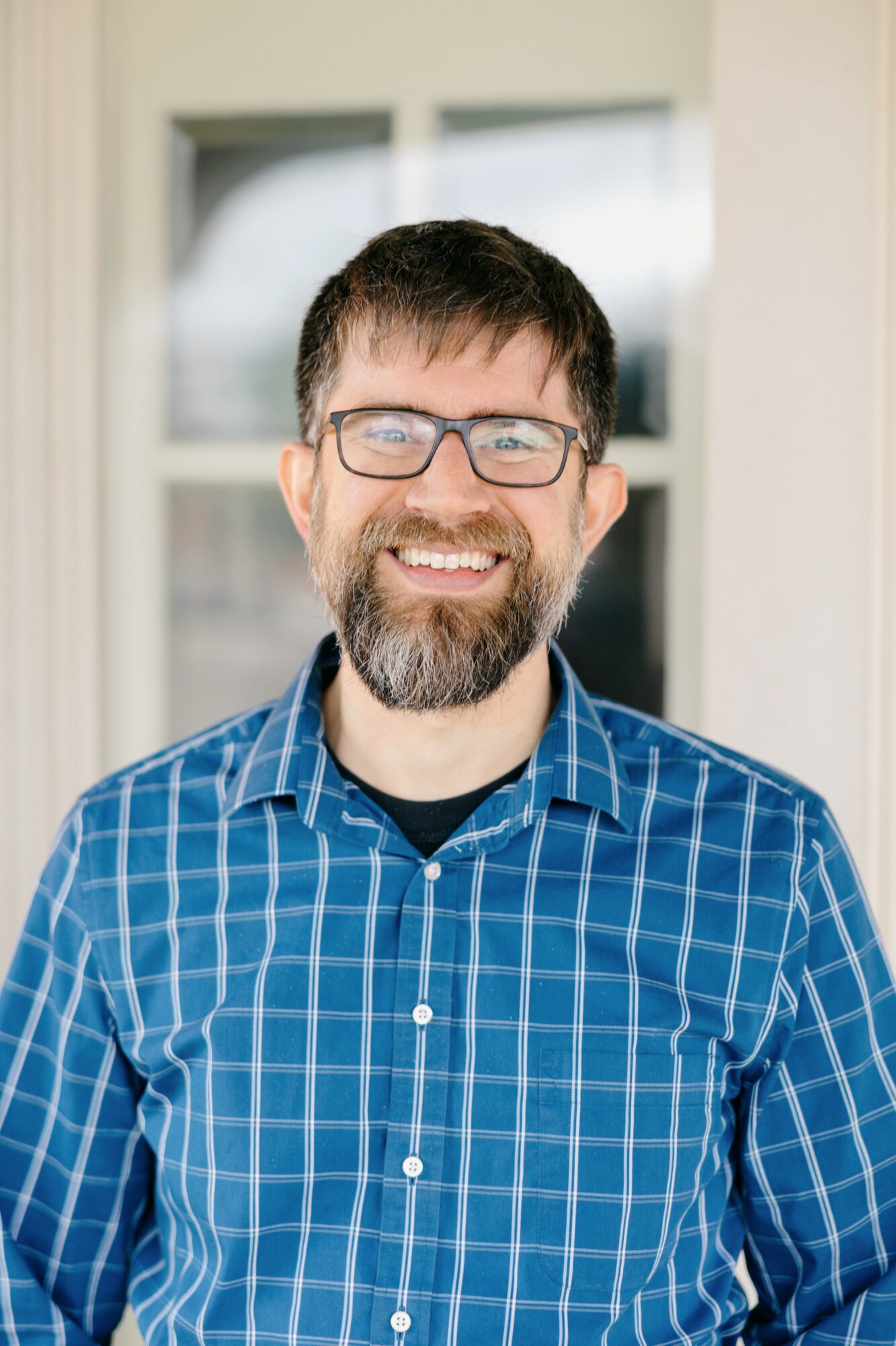 Man with glasses and beard smiling, wearing a blue checkered shirt, standing outside near a window.