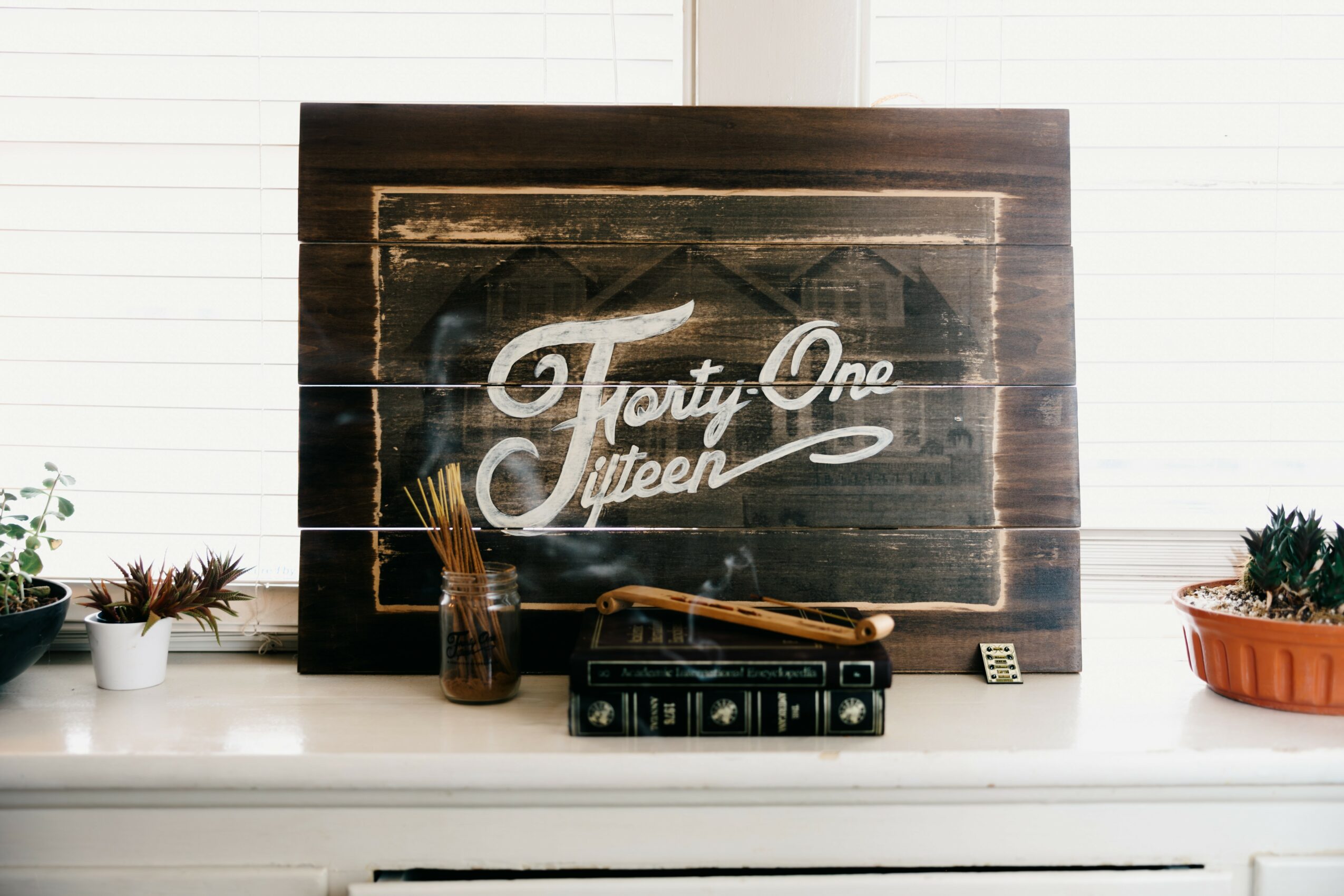 Wooden sign with white cursive text reading 'Forty One Fifteen' on a white surface with plants and objects around.