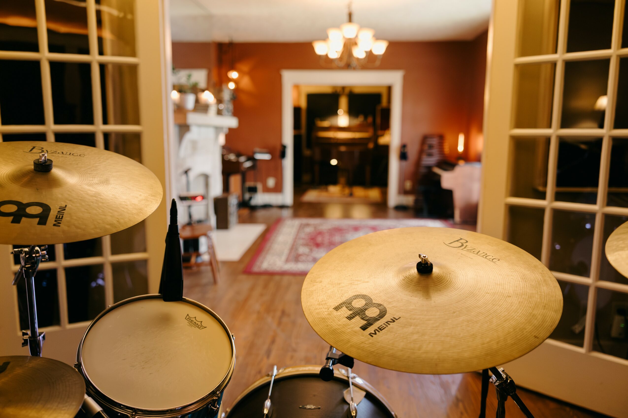 View of a drum set in a room with open French doors, living room with fireplace, and warm lighting.