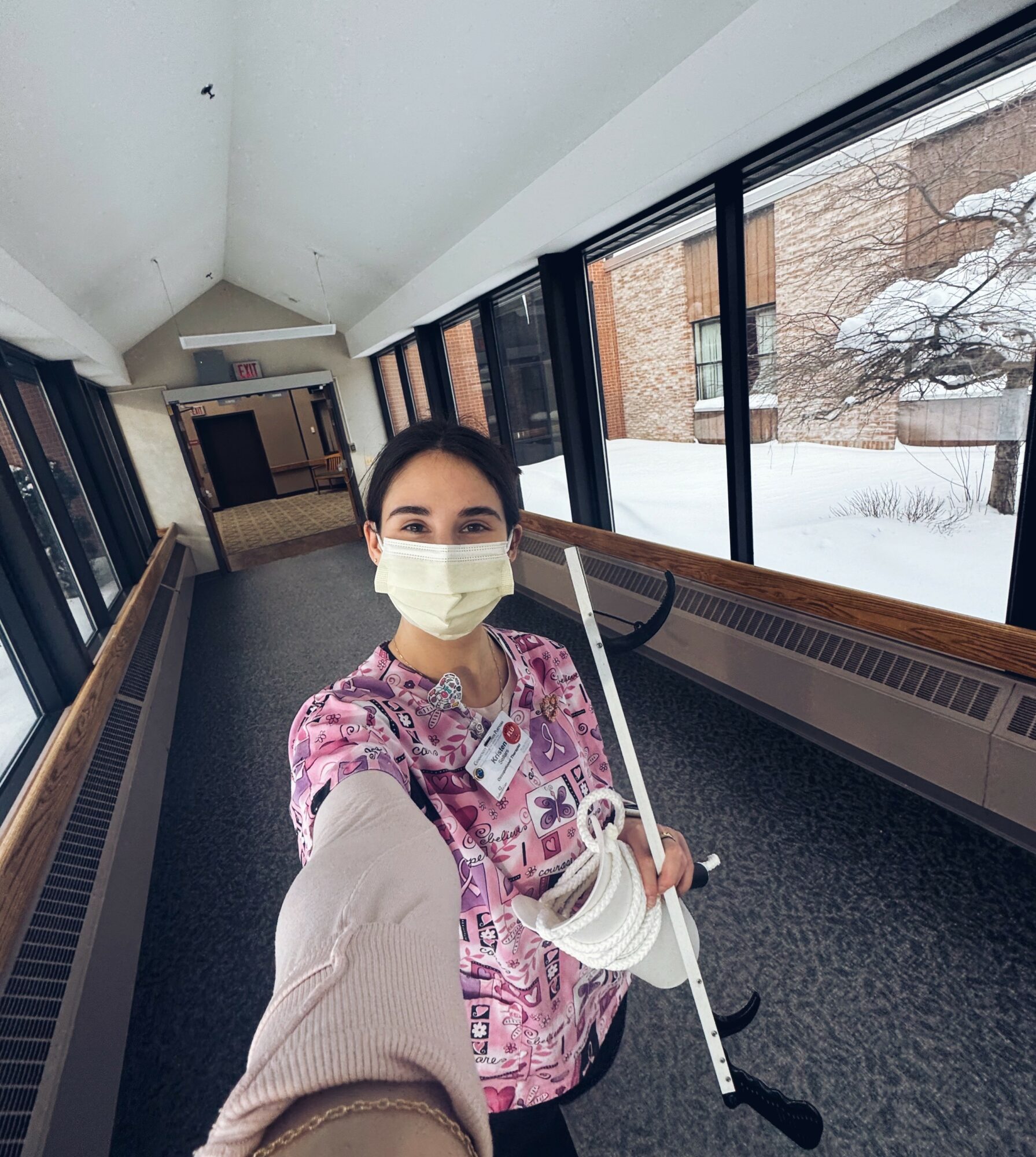 Person wearing a face mask and colorful scrubs taking a selfie in a corridor with large windows and snow outside.