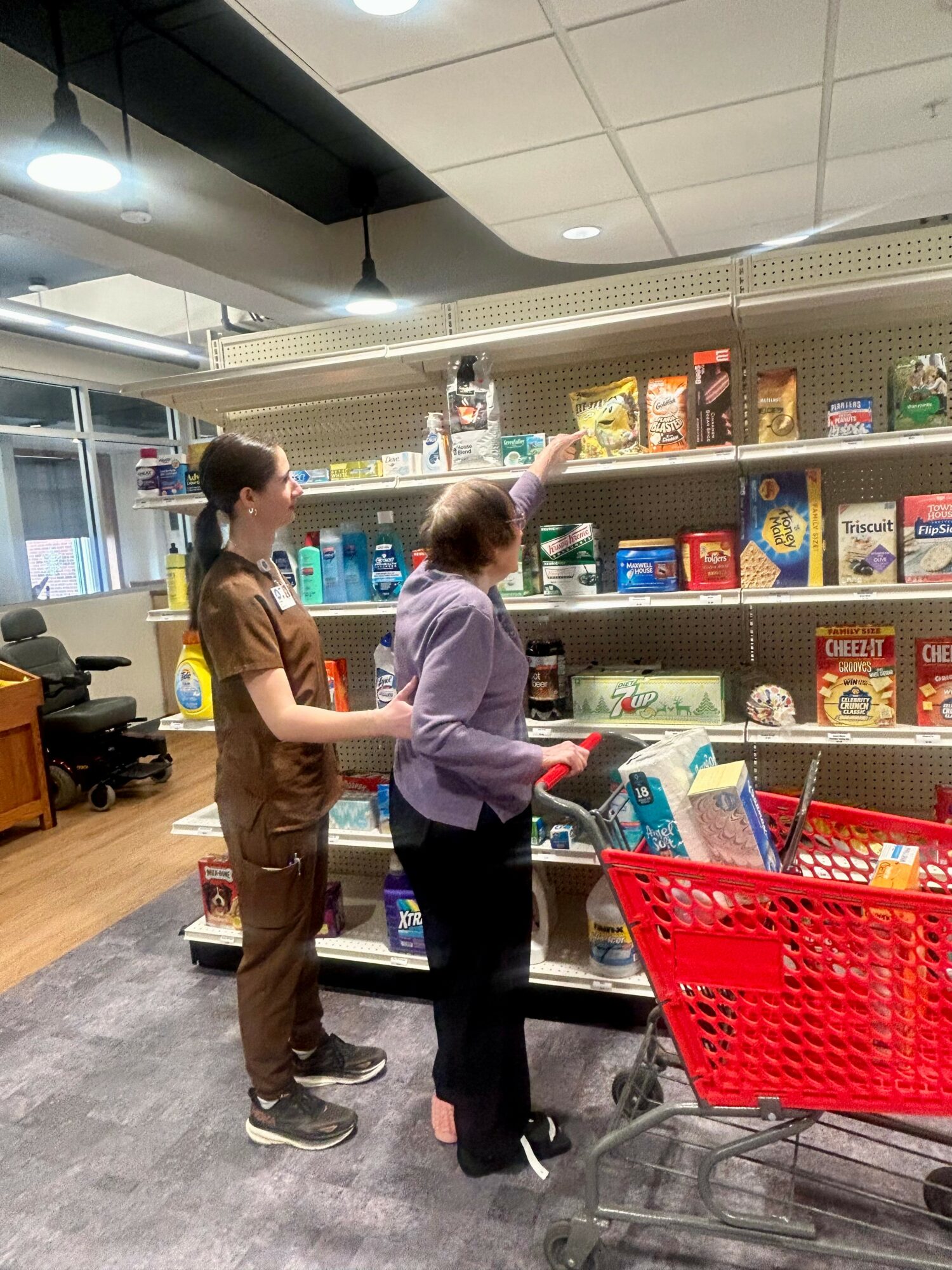Two women shopping in a store aisle with shelves stocked with food and household items, one reaching for an item on the top shelf.