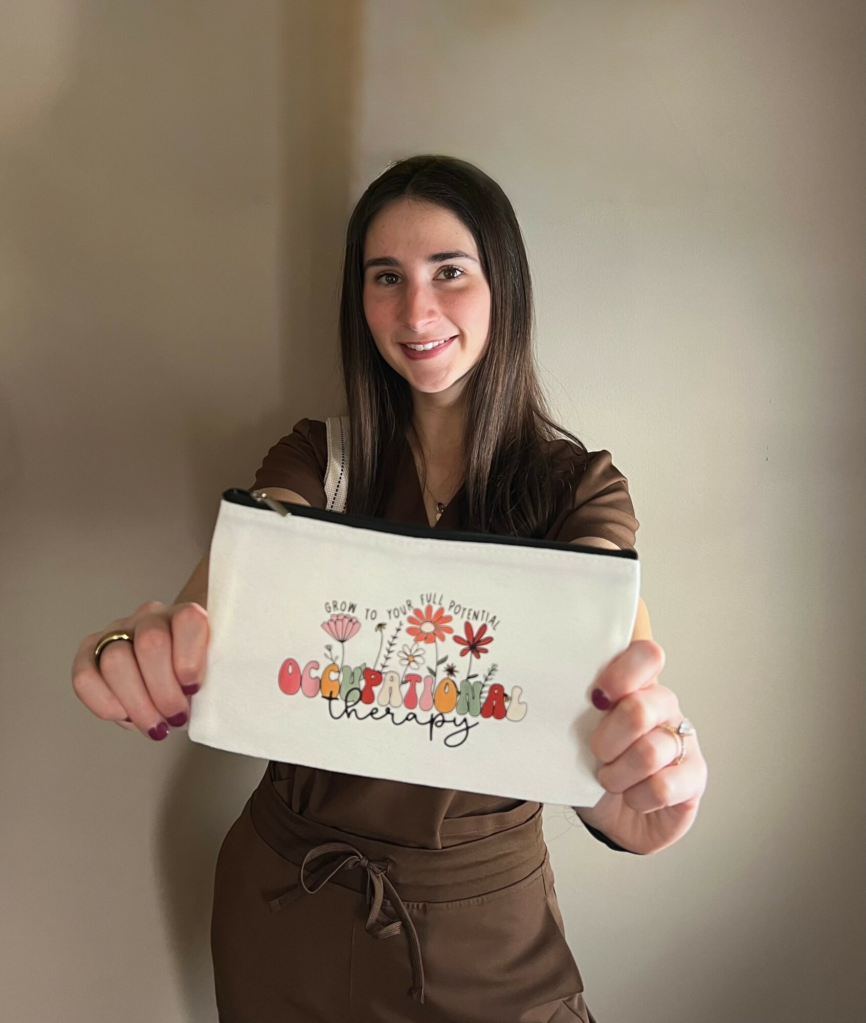 Young woman with long dark hair holding a white pouch with colorful text and flowers, smiling, standing indoors.