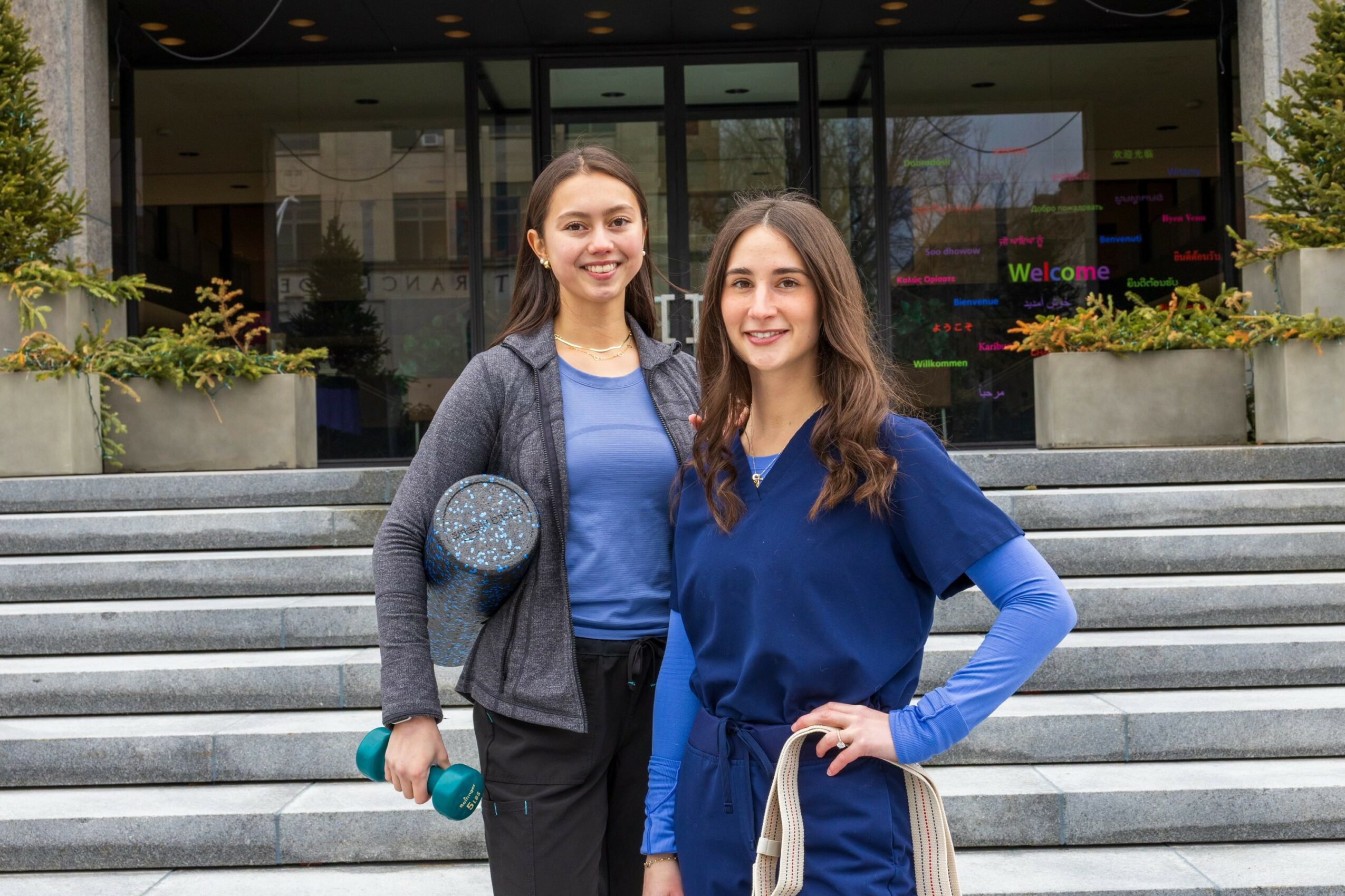 Two young women stand on stairs outside a building, smiling, with glass doors behind them, one holding a water bottle.