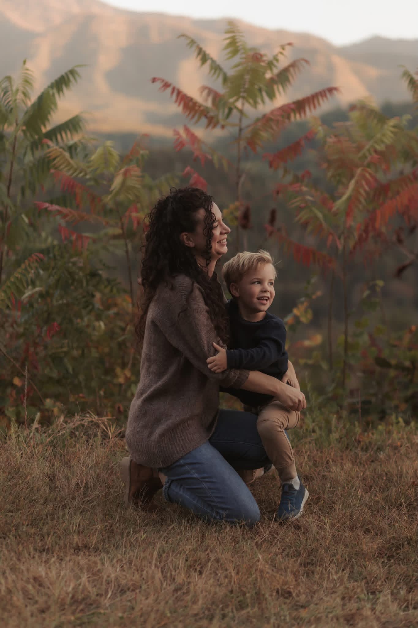 Woman kneeling outdoors with a young child, surrounded by trees and mountains in the background.