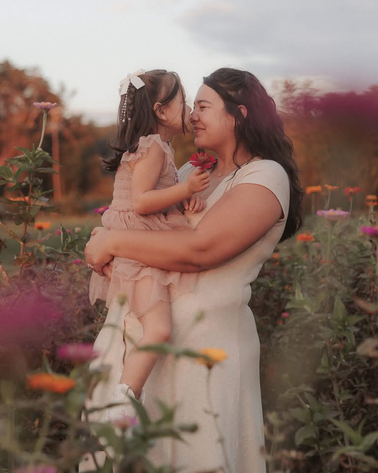 Woman holding a young girl in a flower garden, both smiling and touching noses, surrounded by colorful flowers.