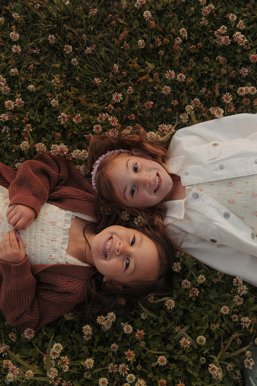Two women lying on grass with small flowers, looking up at the camera, smiling, one wearing a white jacket, the other a brown sweater.