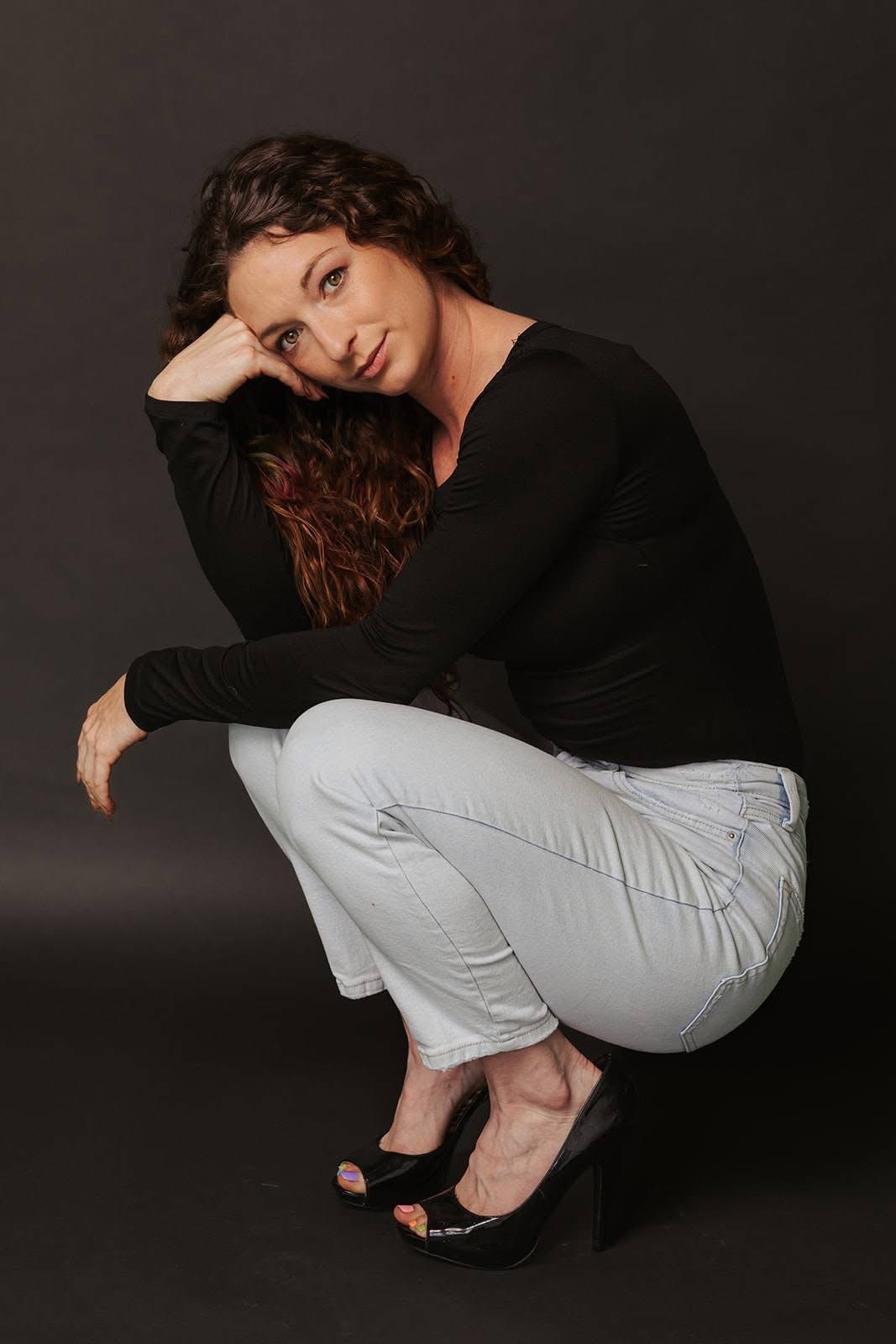 Woman with curly hair in black top and light pants squatting, resting her head on her hand against a dark background.