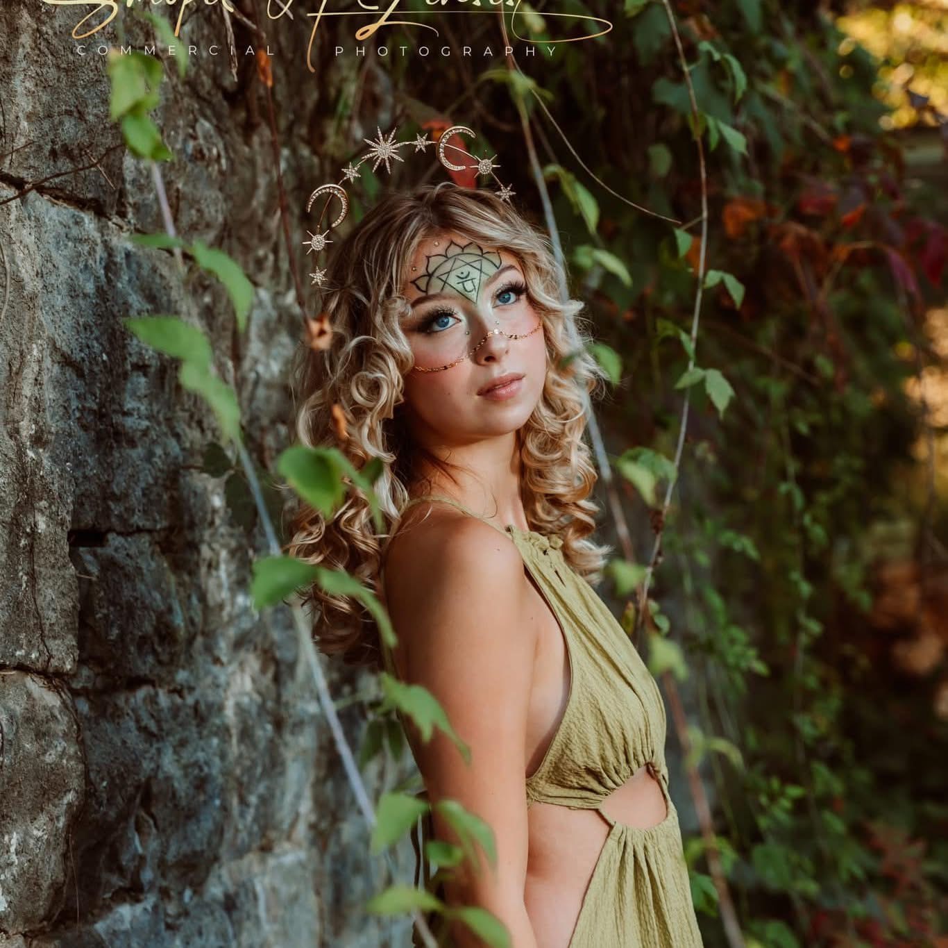 Young woman with curly hair and face paint, wearing a green dress, standing near a stone wall with greenery.