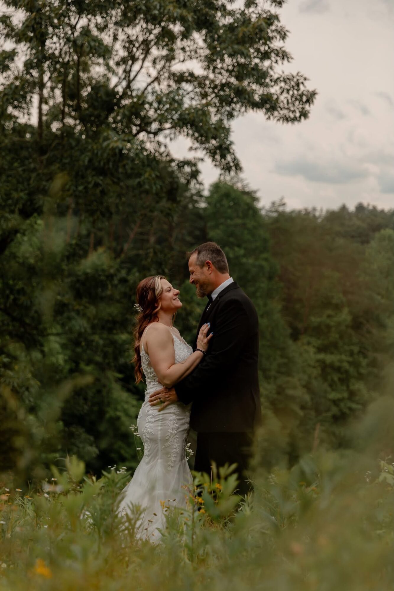 A couple in wedding attire standing outdoors, embracing each other with trees and sky in the background.