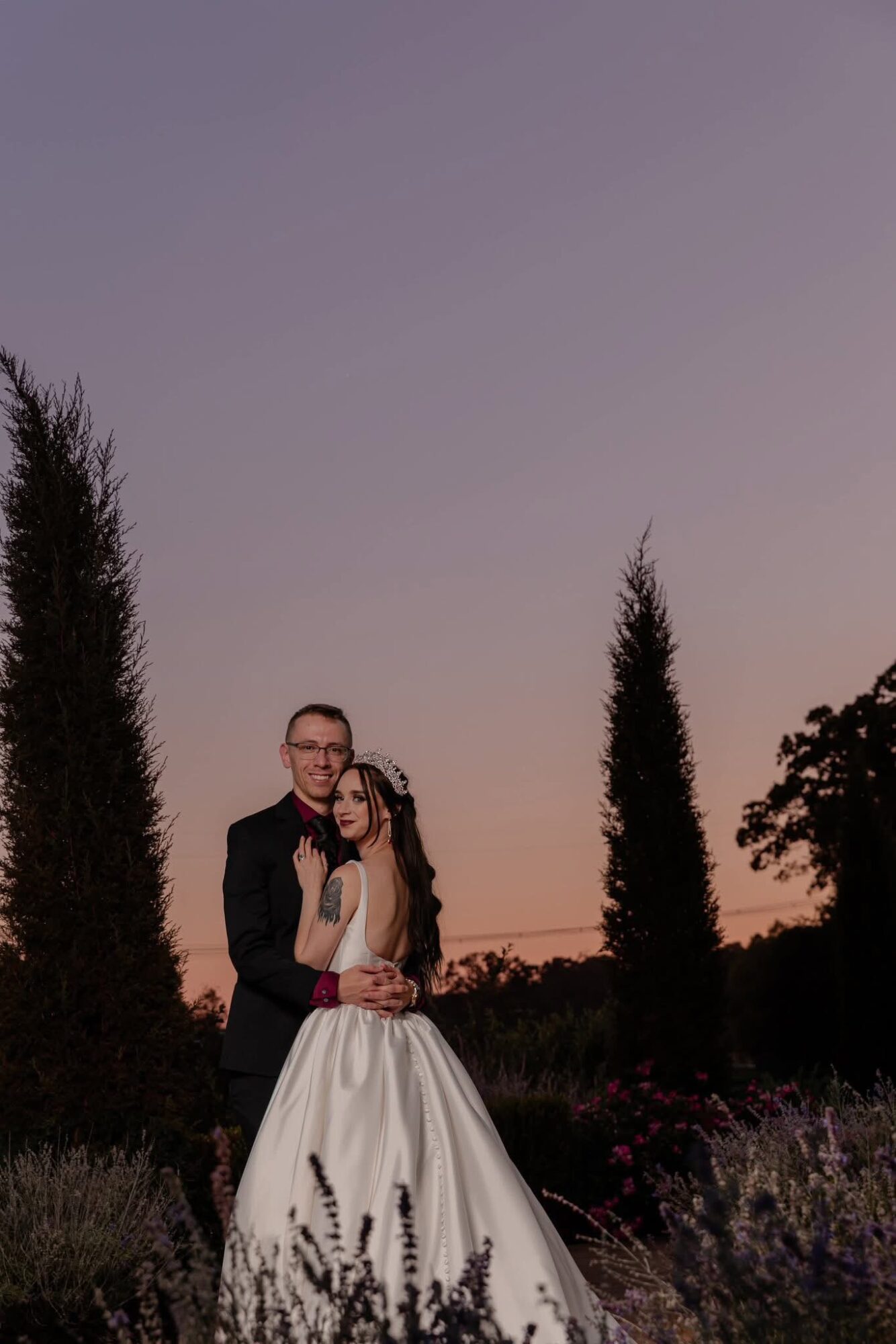 A couple in formal attire standing outdoors during sunset, with trees and a purple sky in the background.