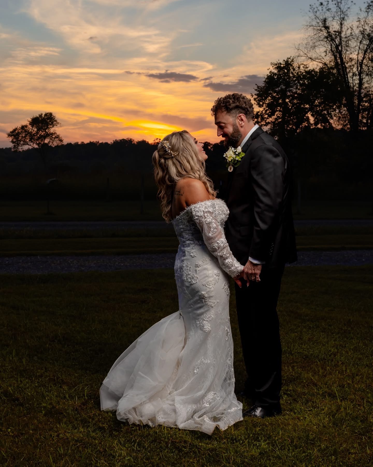 Bride and groom holding hands and smiling outdoors at sunset, with trees and a colorful sky in the background.