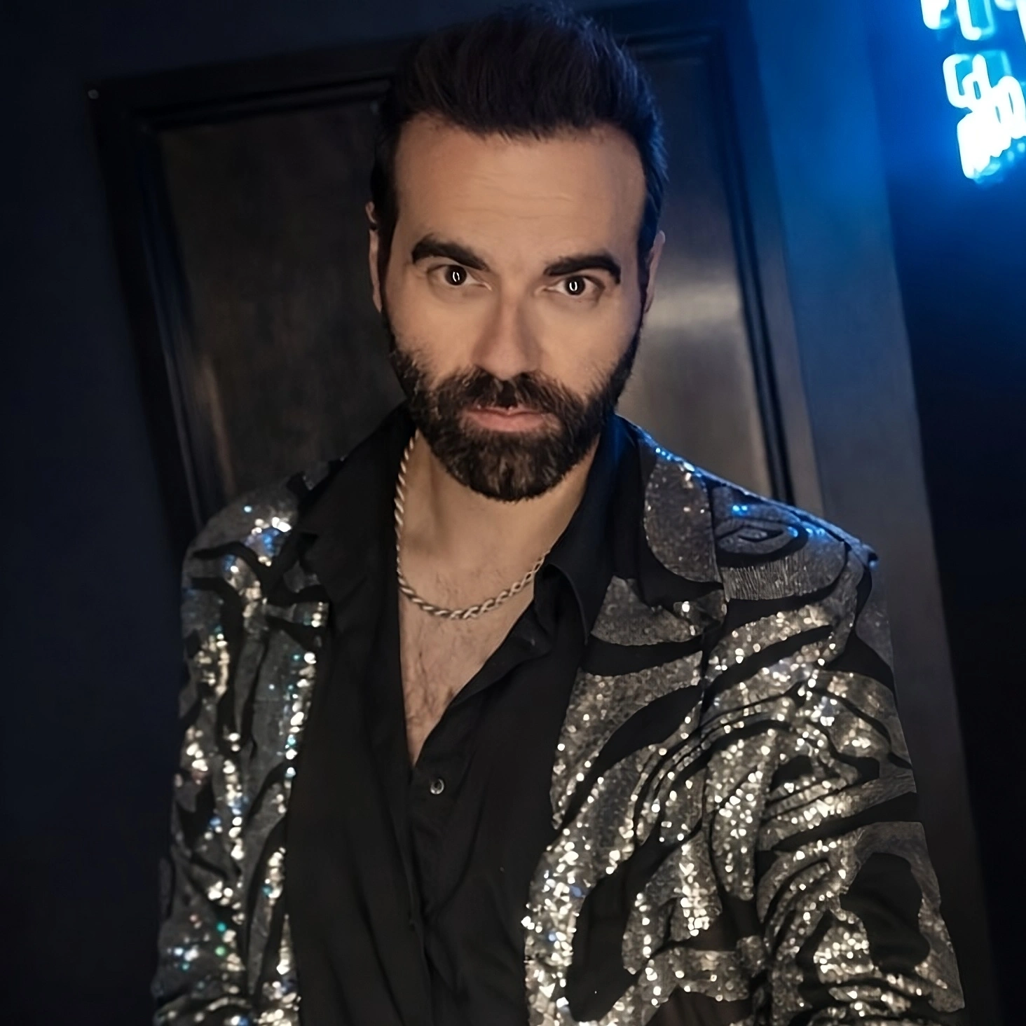 Man with dark hair and beard wearing a shiny, patterned jacket and black shirt, standing indoors with blue lighting.
