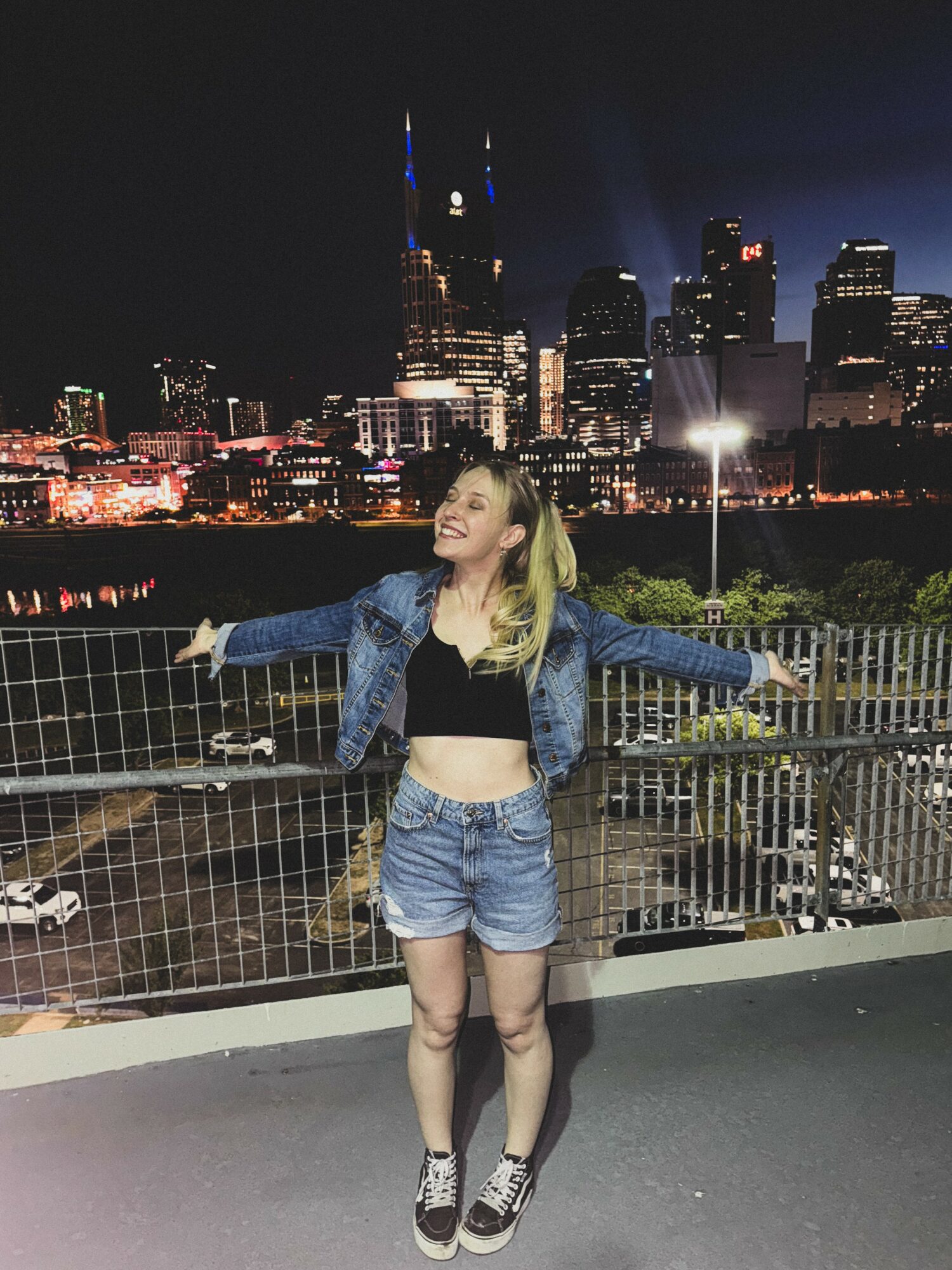 Young woman with blonde hair stands on a rooftop at night, arms outstretched, city skyline with lit buildings behind her.
