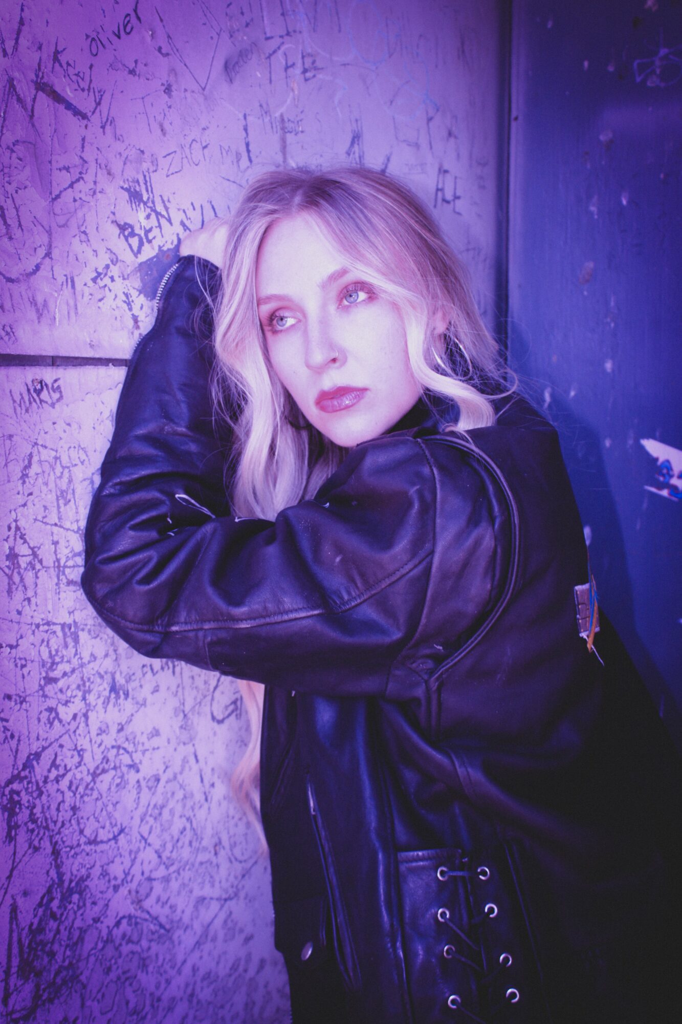 Young woman with blonde hair leaning against graffiti-covered wall, wearing a black leather jacket, looking to the side.