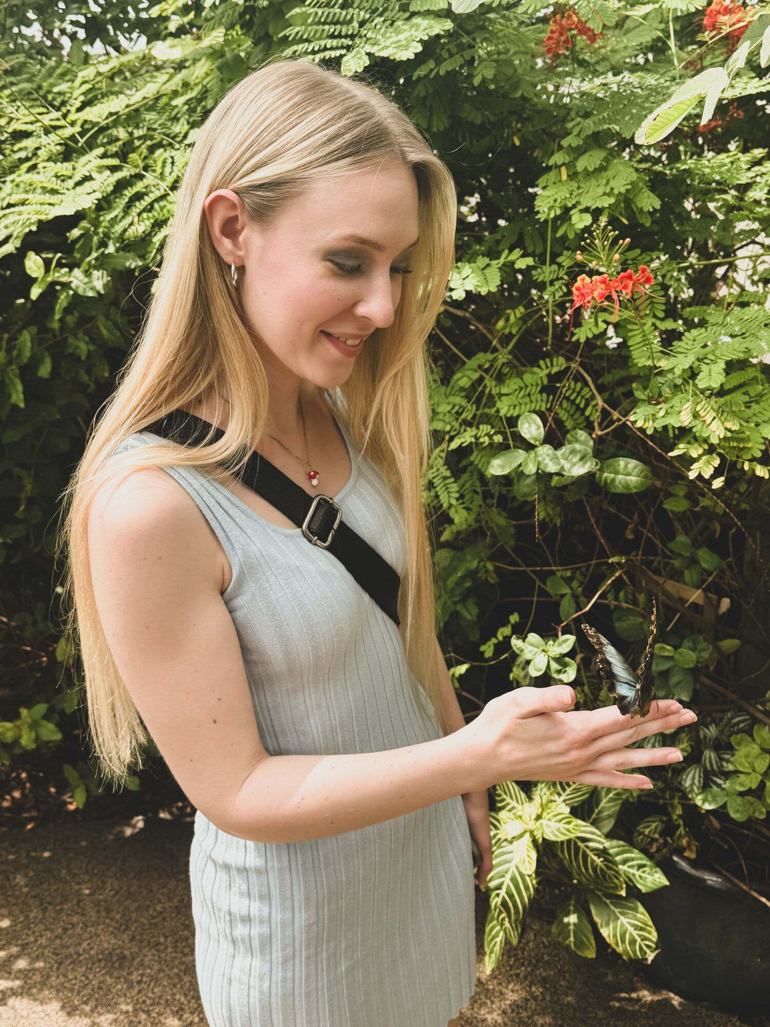 Young woman with long blonde hair smiling outdoors, holding a butterfly on her hand, surrounded by green plants.
