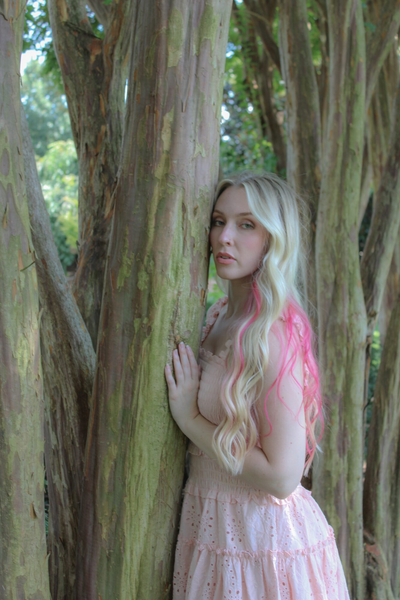 Young woman with long blonde hair in a pink dress stands among trees, touching a tree trunk, in a forest setting.