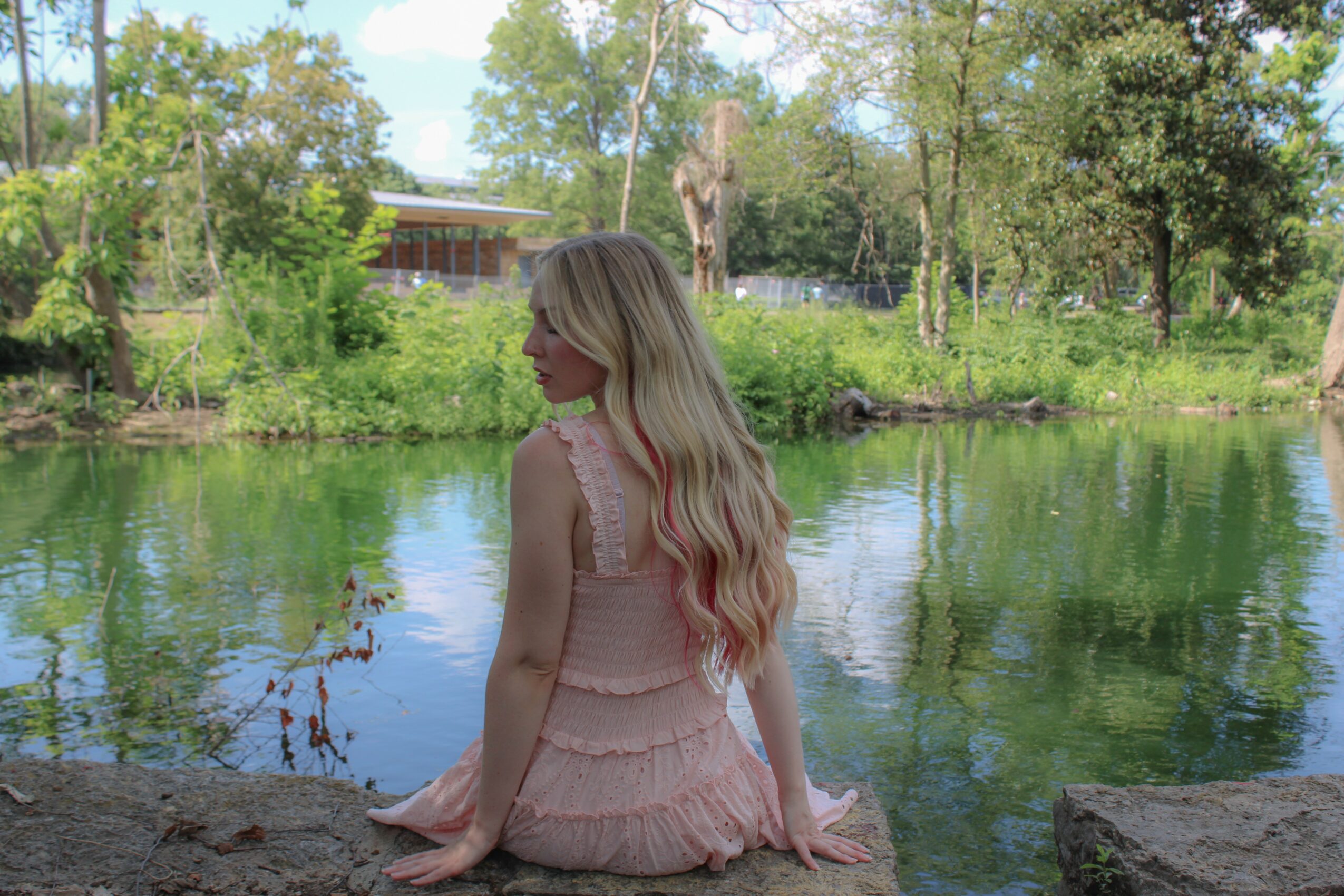 Woman with long blonde hair sitting by a river, surrounded by trees and greenery, facing sideways.