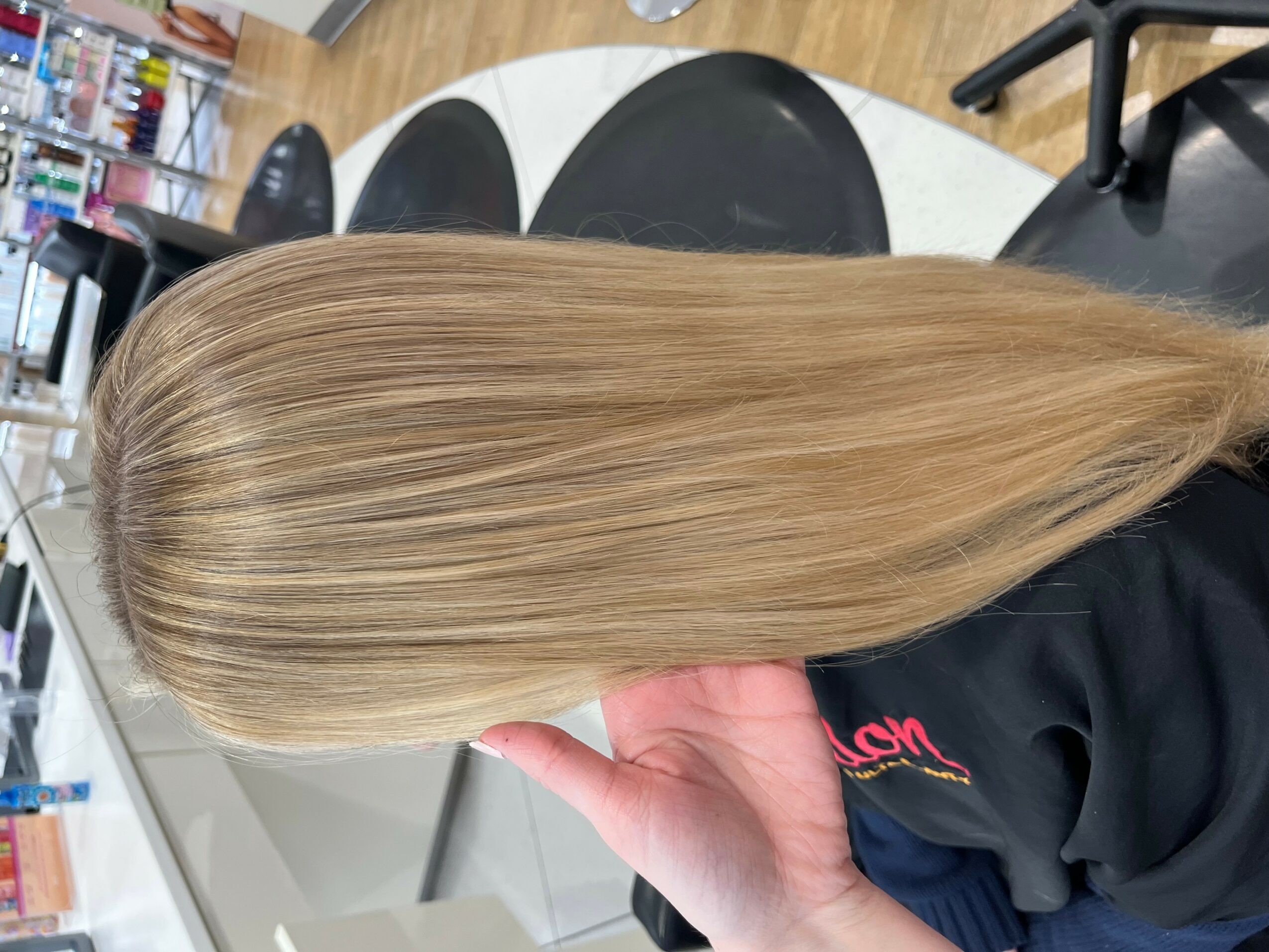 Person with long, straight, blonde hair in a salon chair, holding a section of hair with their hand.