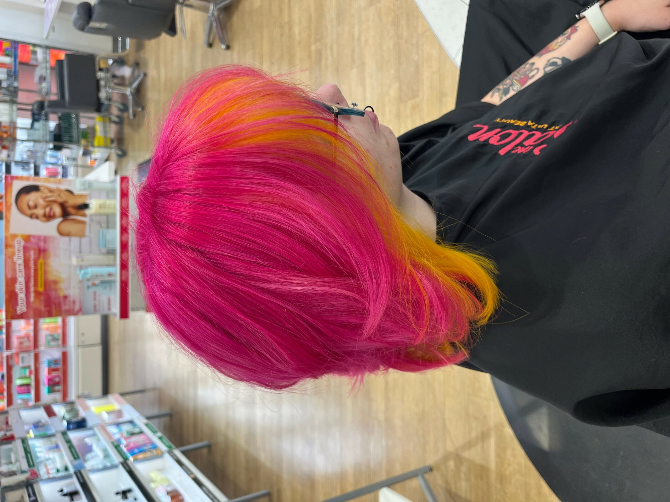 Side view of a person with vibrant pink and yellow hair in a salon, wearing a black cape, with salon equipment in background.