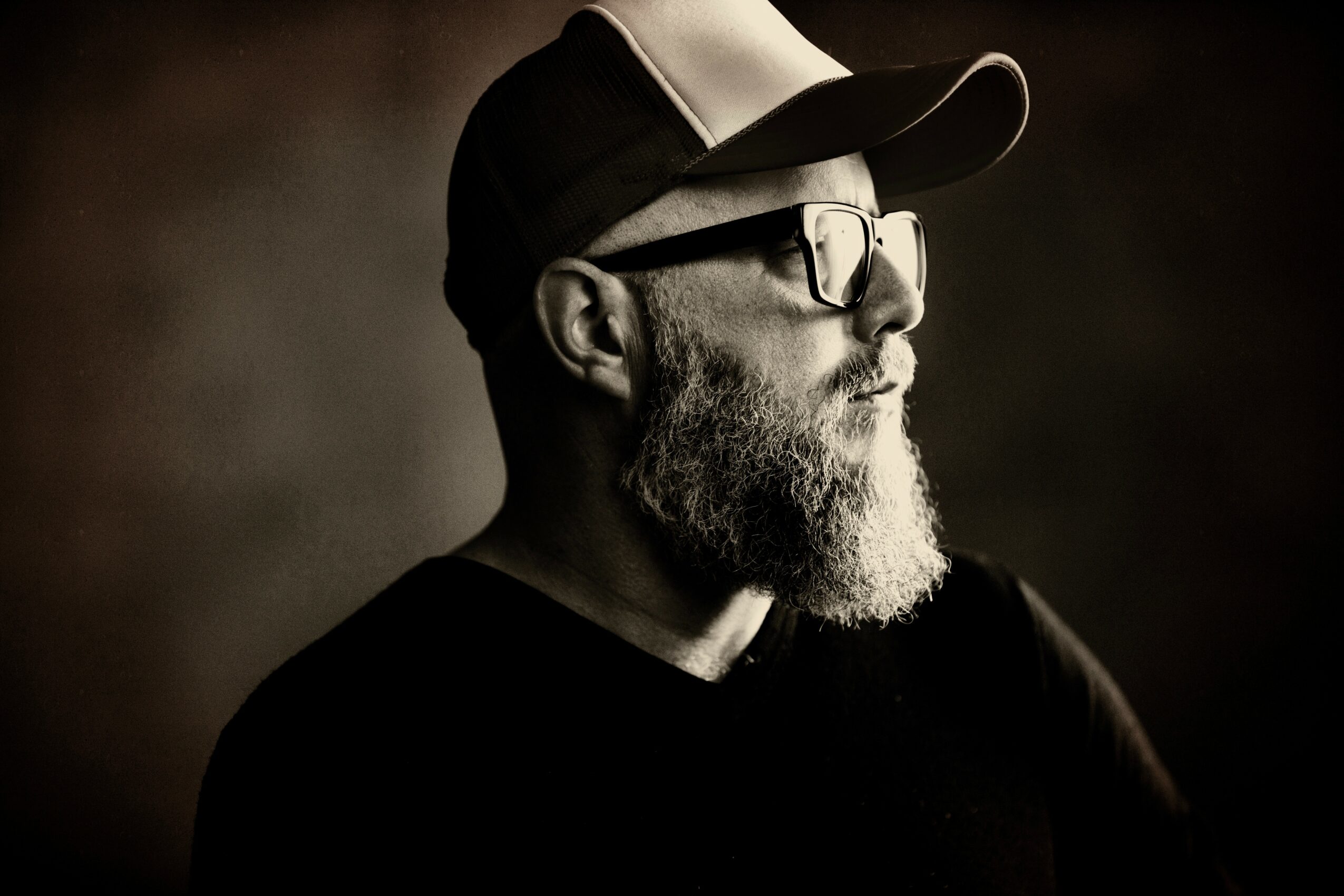 A bearded man wearing glasses and a baseball cap, looking to the right, in black and white.