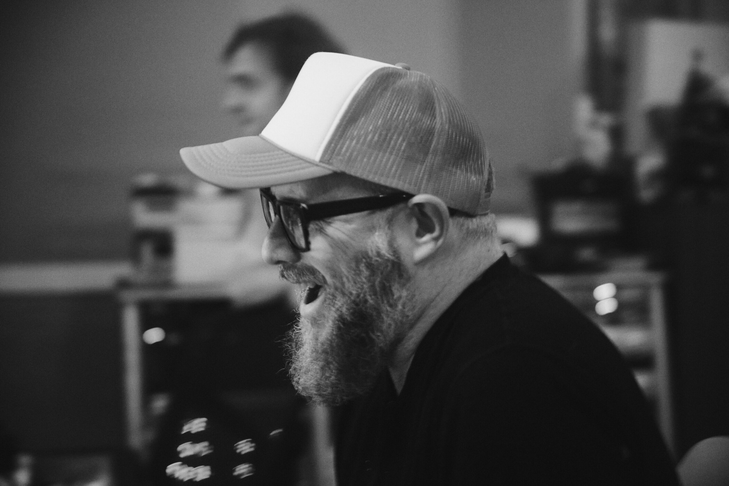 Man with glasses and a beard wearing a cap, smiling, in a room with blurred background.
