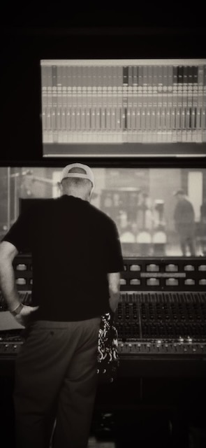 Person wearing a cap and black shirt standing in front of audio mixing console, with a large screen displaying waveforms above.