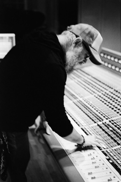 Person working at a large audio mixing console in a recording studio, wearing a cap and hoodie.