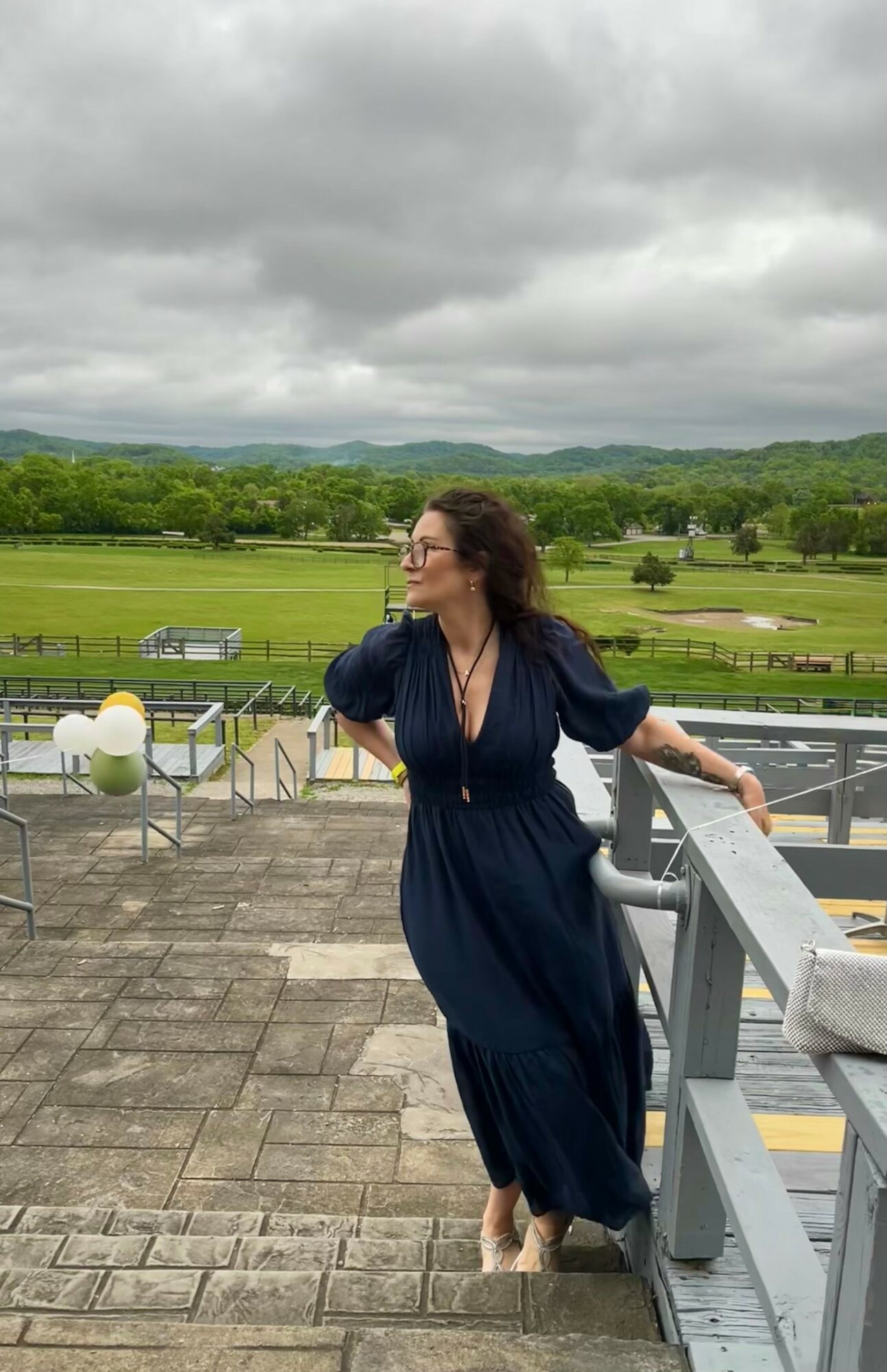 Woman in a long dark dress standing on a balcony with a landscape of green fields and cloudy sky behind her.