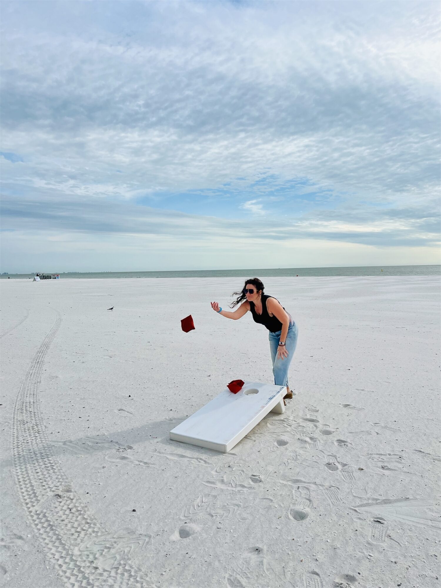 Person in swimsuit throws red objects on a white platform in a sandy beach with blue sky and clouds