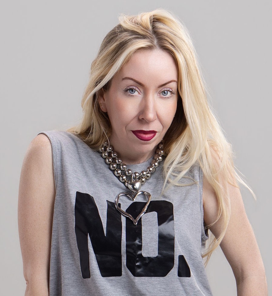 Woman with blonde hair wearing a sleeveless gray top with bold black letters, a pearl necklace, and dark lipstick, looking at the camera.