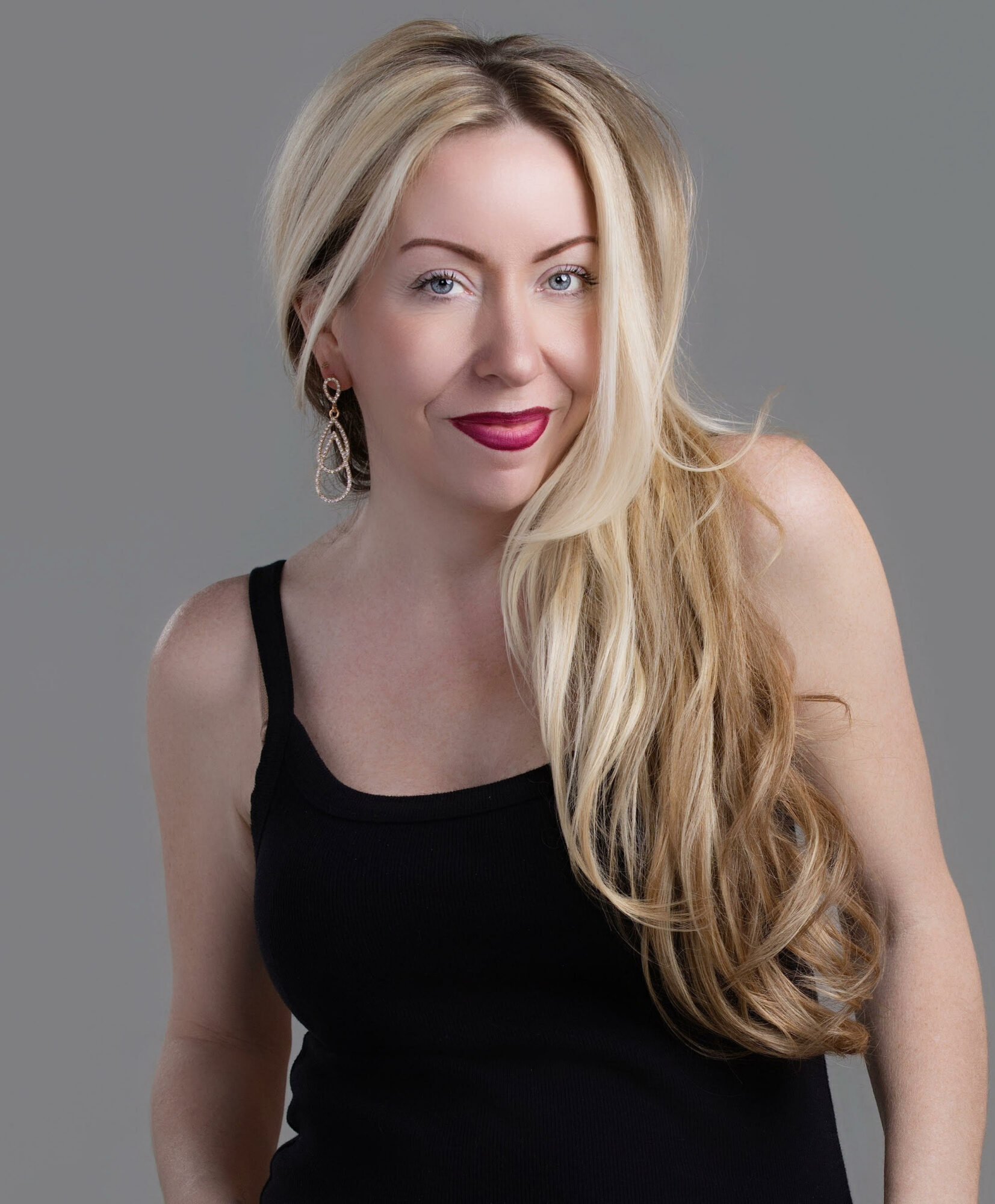 Woman with long blonde hair wearing a black top, smiling, against a gray background.