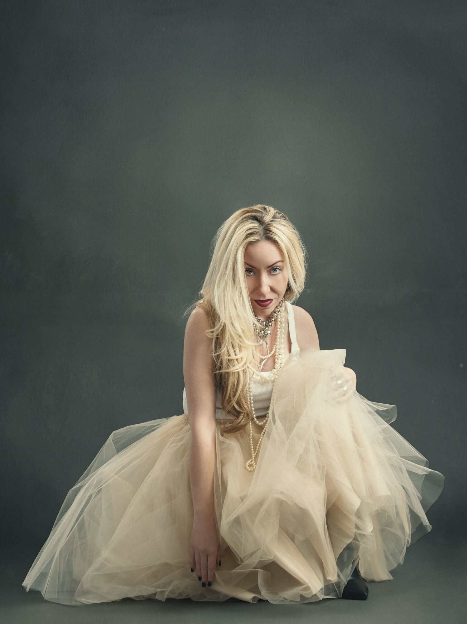 Woman with long blonde hair in a beige dress and tulle skirt, sitting on the floor against a dark background.