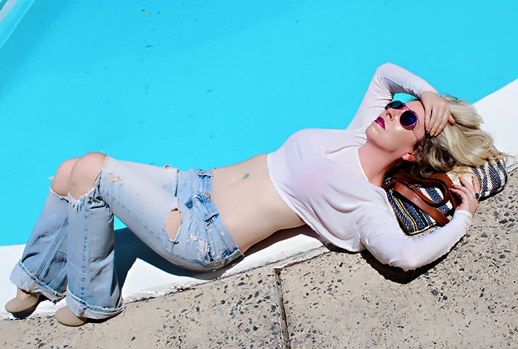 Woman lying on poolside with sunglasses, white top, and ripped jeans, relaxing with one arm behind her head.