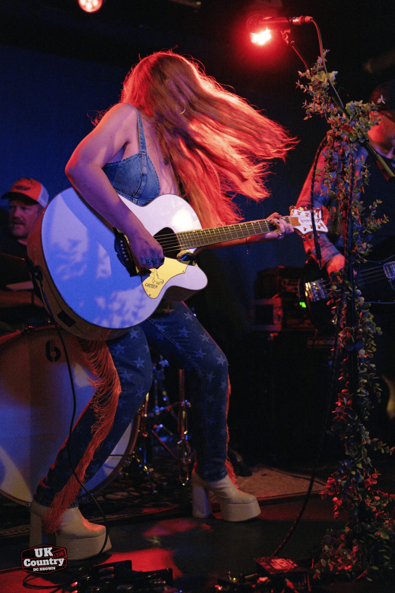 Woman with long hair playing an acoustic guitar on stage, illuminated by red and blue lights, with a floral decoration nearby.