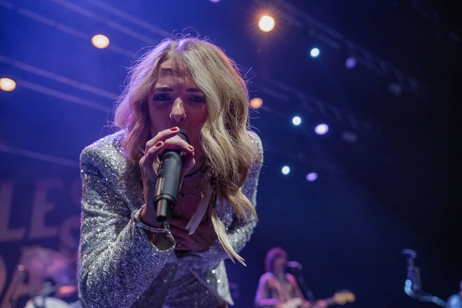 A woman with blonde hair singing into a microphone on stage with colorful lights in the background.