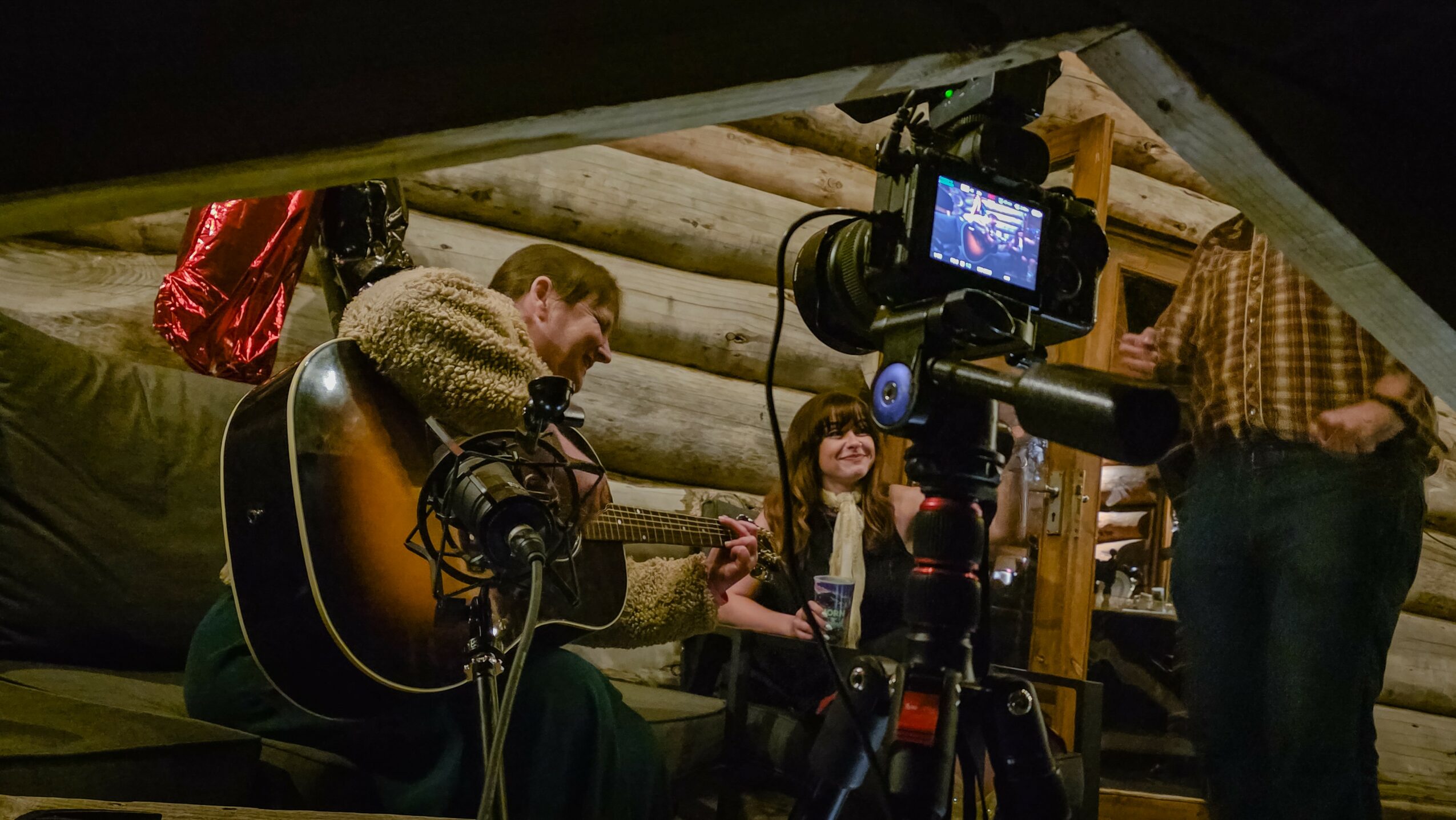 Person playing guitar in a cozy, log cabin setting with camera equipment recording them, two other people nearby, warm lighting.
