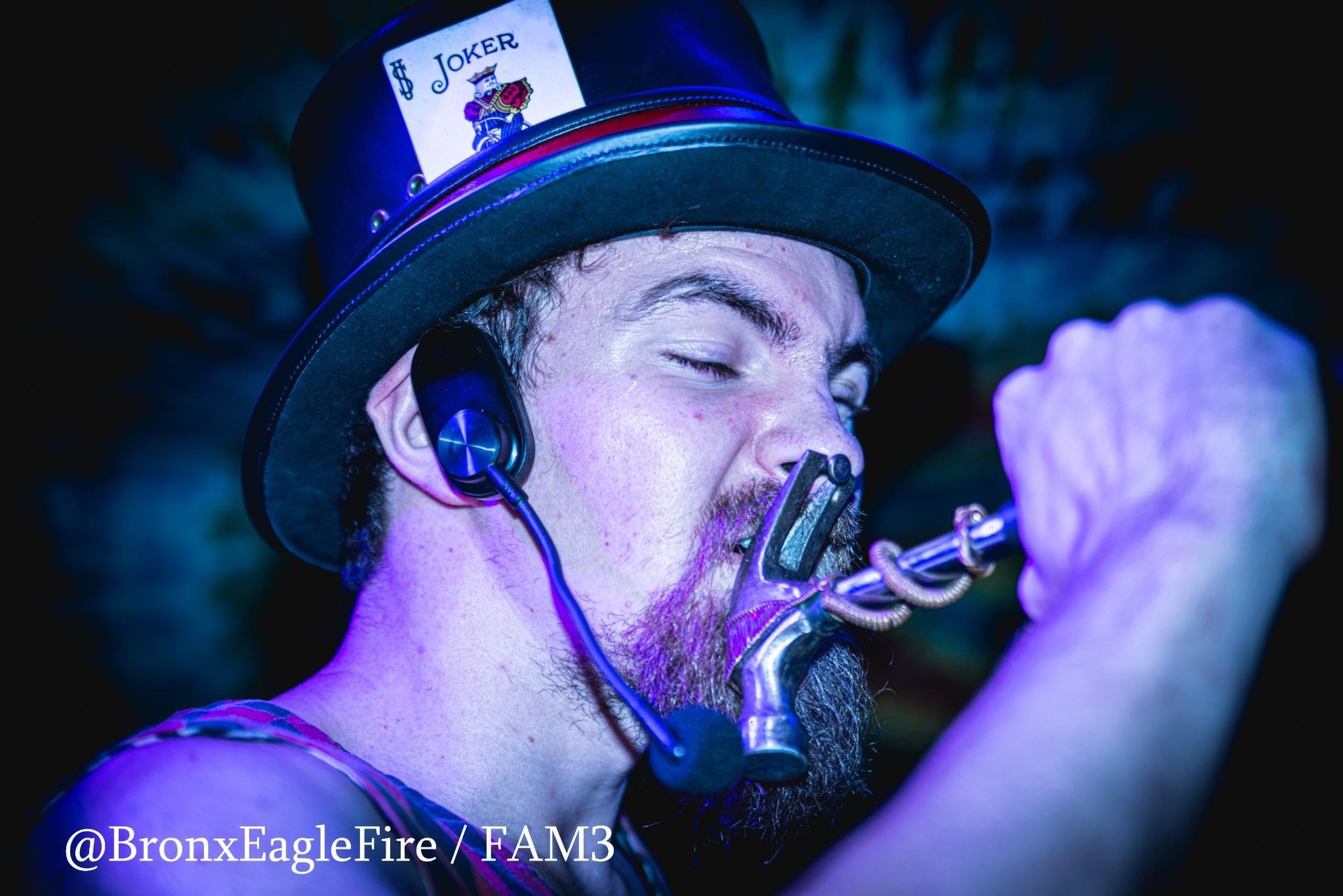 Person wearing a top hat with a Joker card, headphones, and a chain in mouth, making a fierce expression.