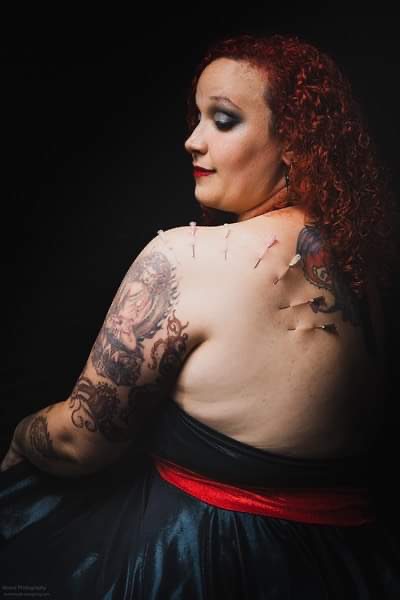 Woman with curly red hair and tattoos on her arm, looking to the side against a dark background.