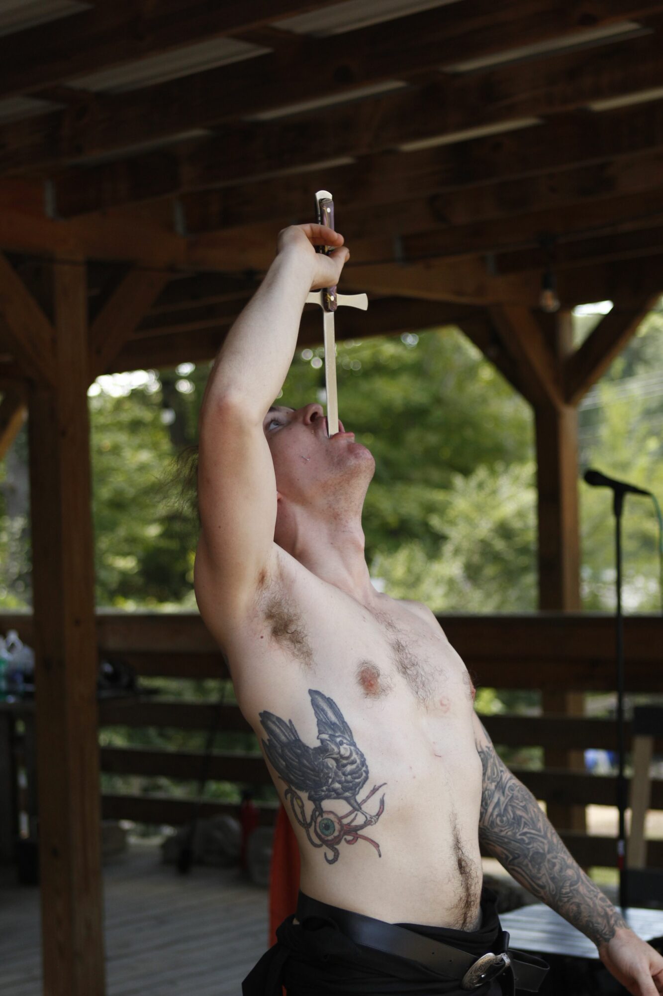 A shirtless person with tattoos on their chest and arm, holding a cigarette, standing outdoors with trees in the background.