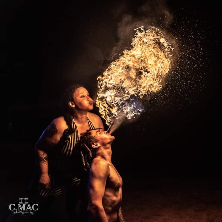 Two women perform a fire-breathing act, with one exhaling a large flame into the air, against a dark background.