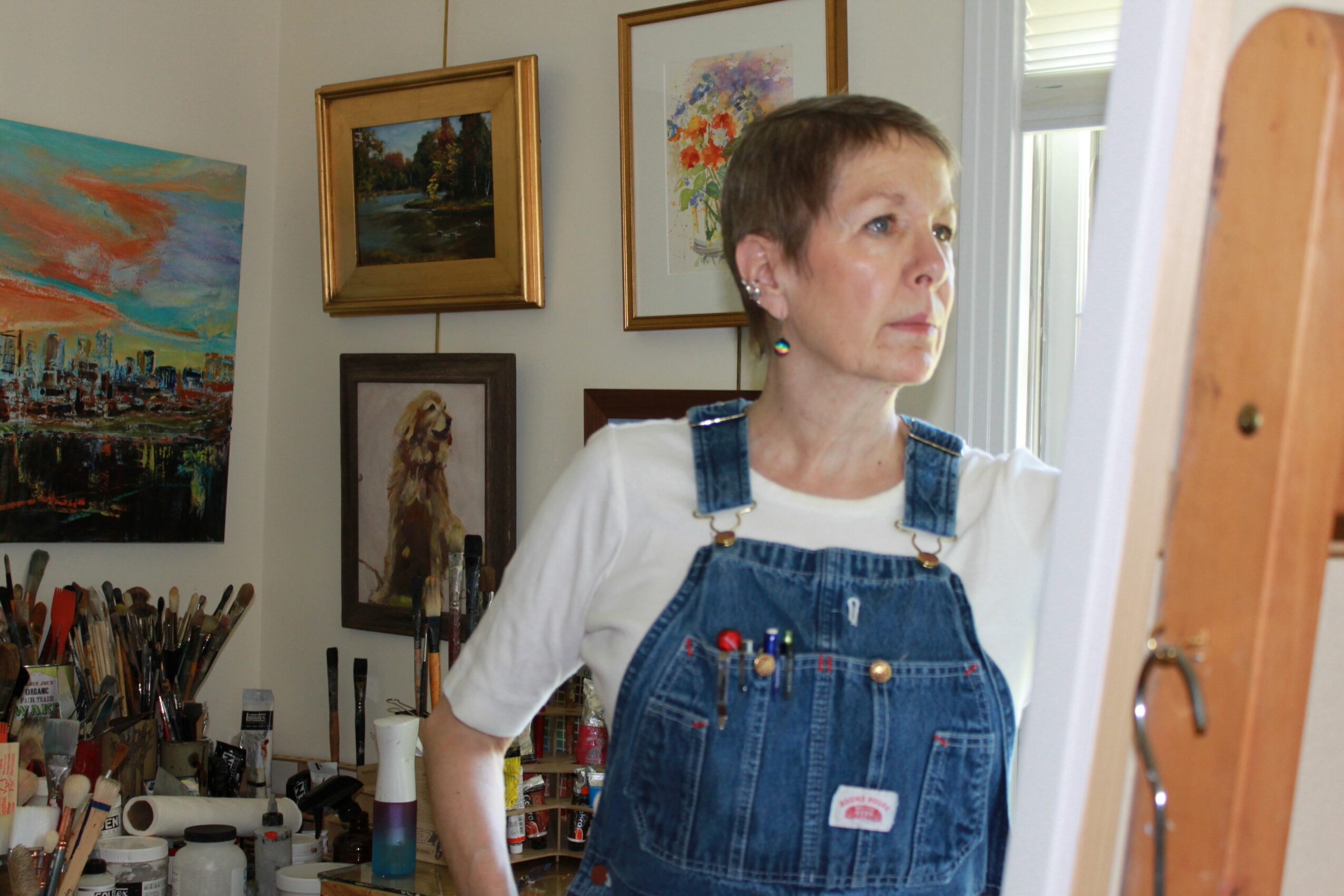 Woman with short hair wearing white shirt and denim overalls looking to the side in an art studio, surrounded by paintings and supplies.