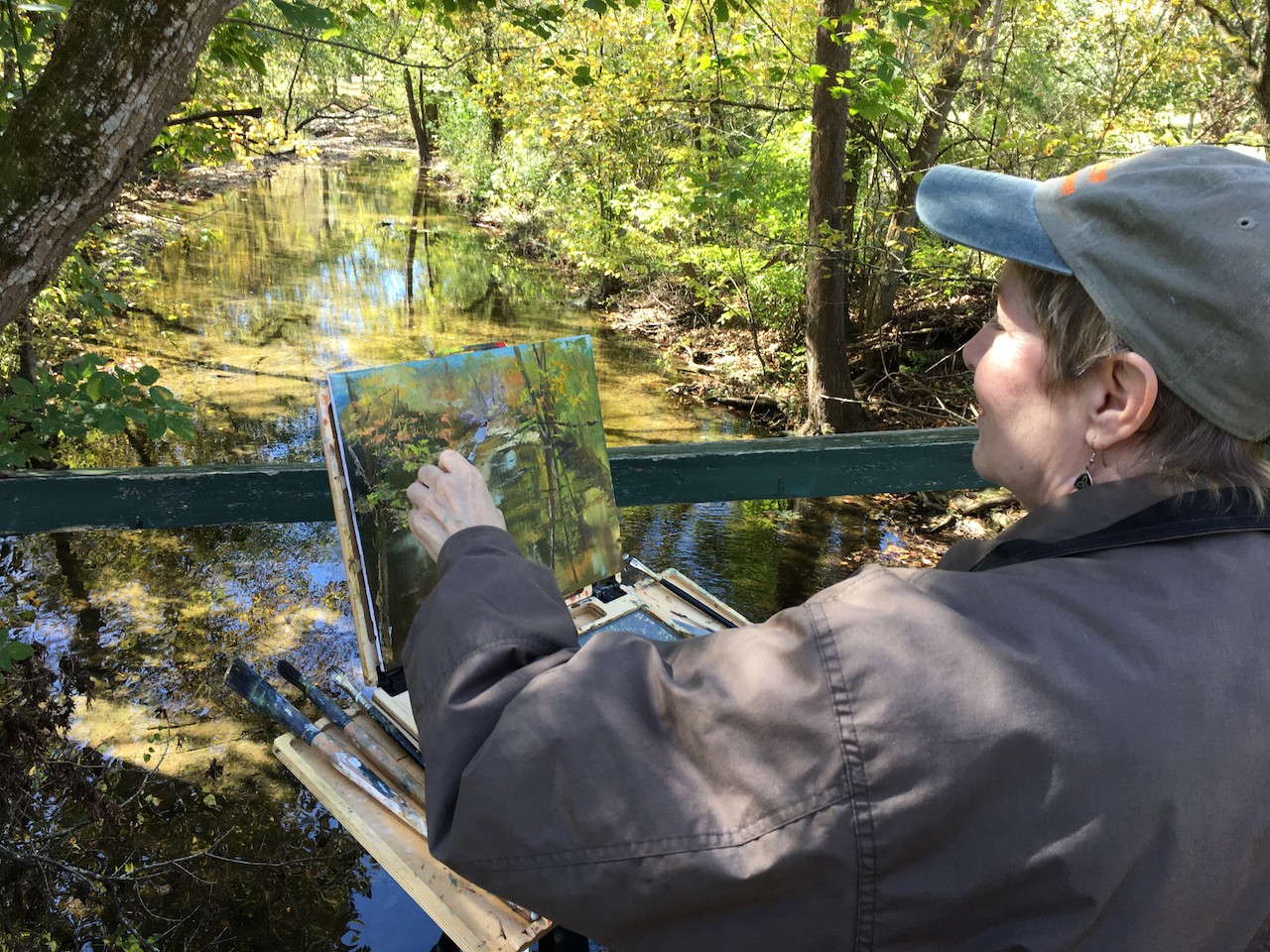 Person painting outdoors near a stream with trees and sunlight, wearing a cap and jacket, holding a paintbrush and palette.