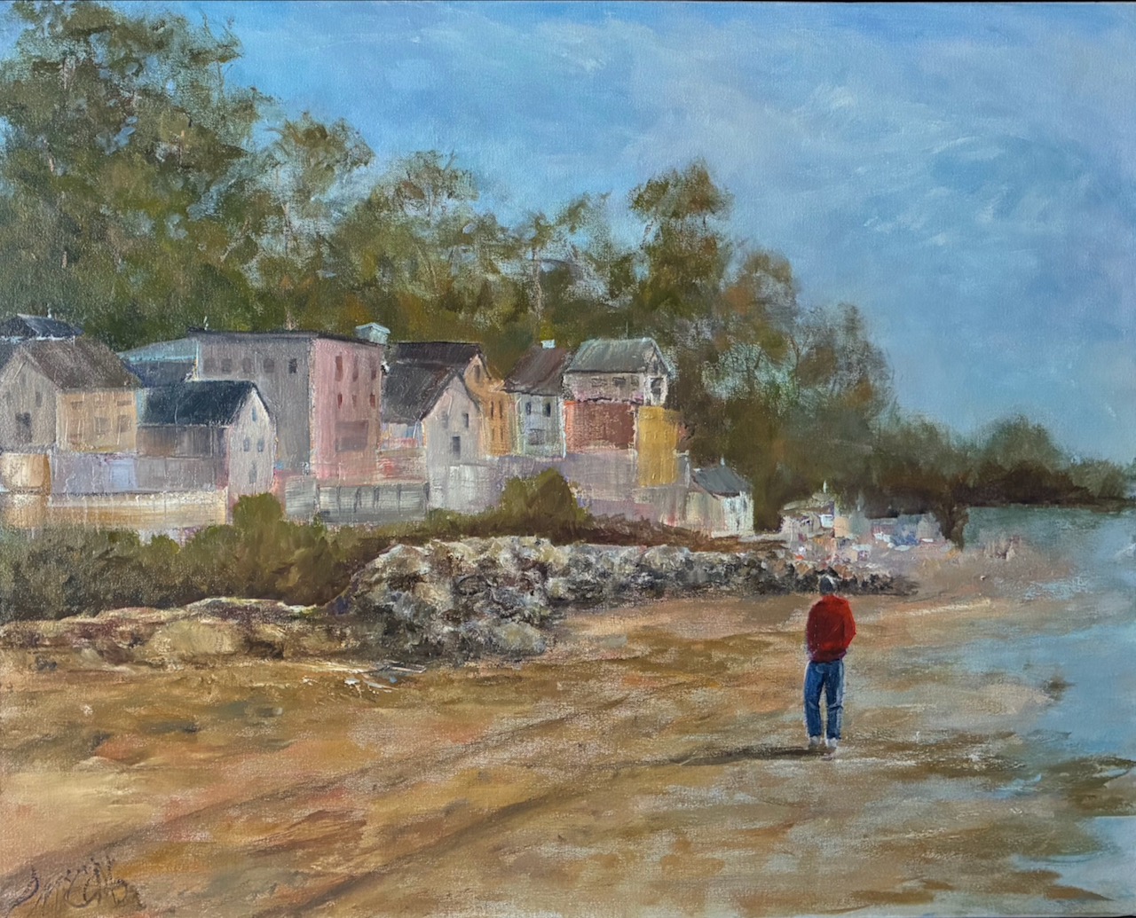 Person standing on a sandy beach near water with colorful houses and trees in the background, under a blue sky.