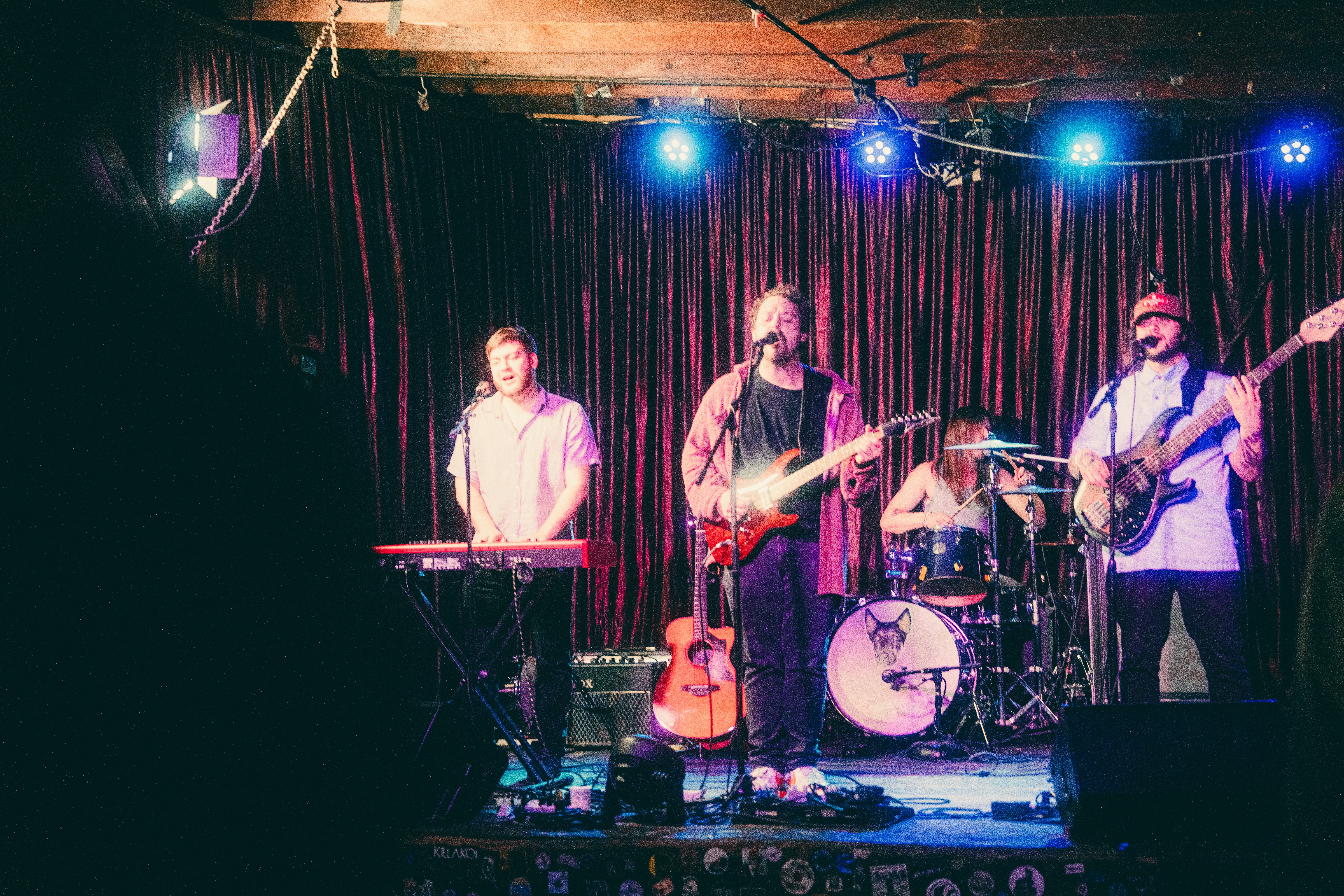 Four musicians perform on stage with a dark curtain background and blue stage lights, one playing keyboard, others with guitars and drums.