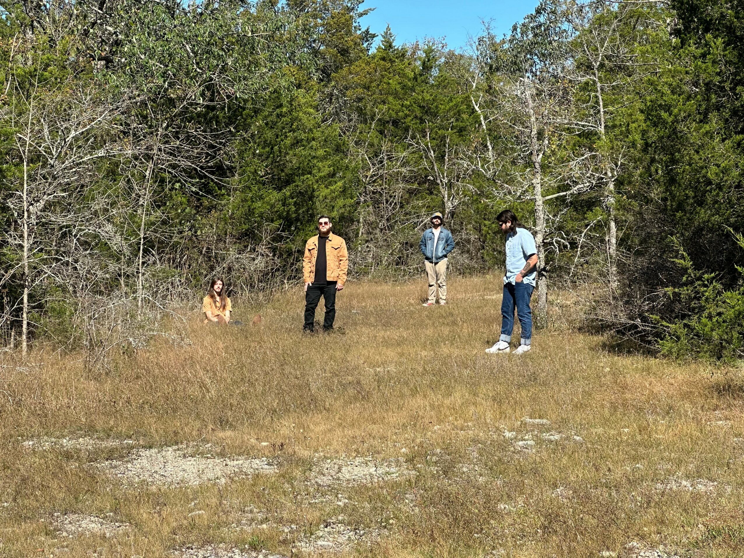 Four people and a dog in a grassy clearing surrounded by trees, with clear sky above.