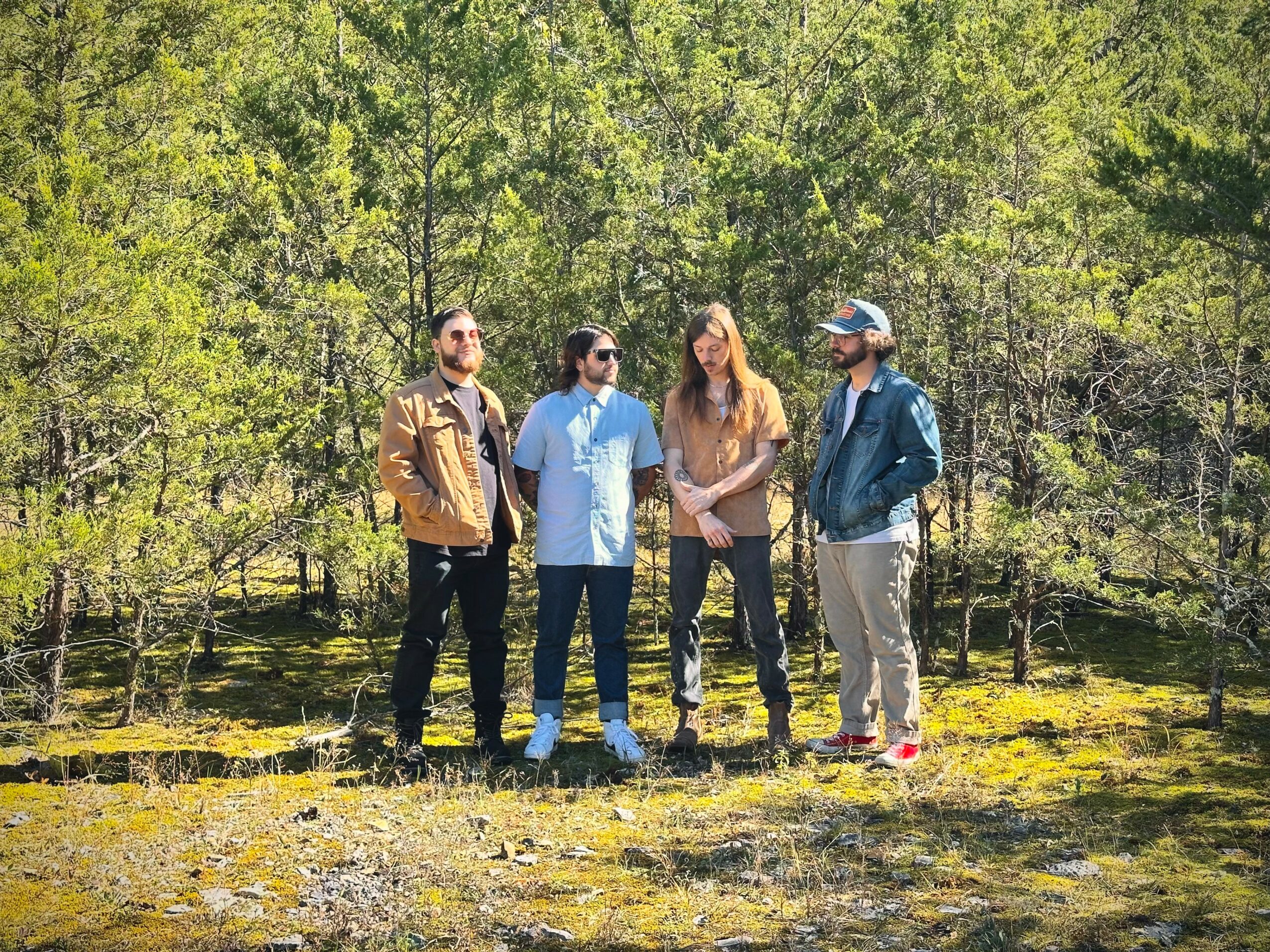 Four people standing outdoors on grass with trees in the background, engaging in conversation.