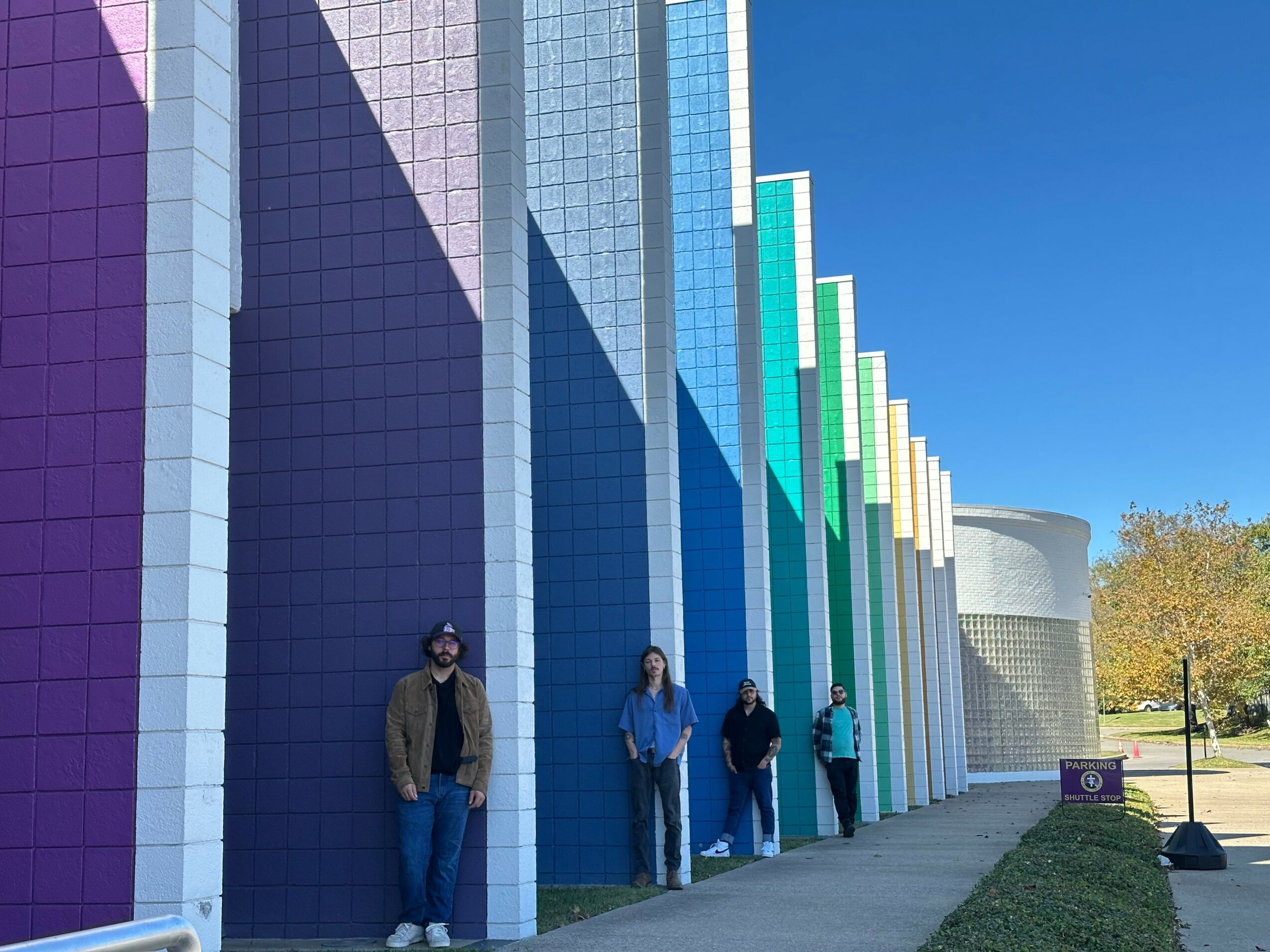 Four people standing in front of a colorful, tall building with vertical rainbow-colored panels, under a clear blue sky.