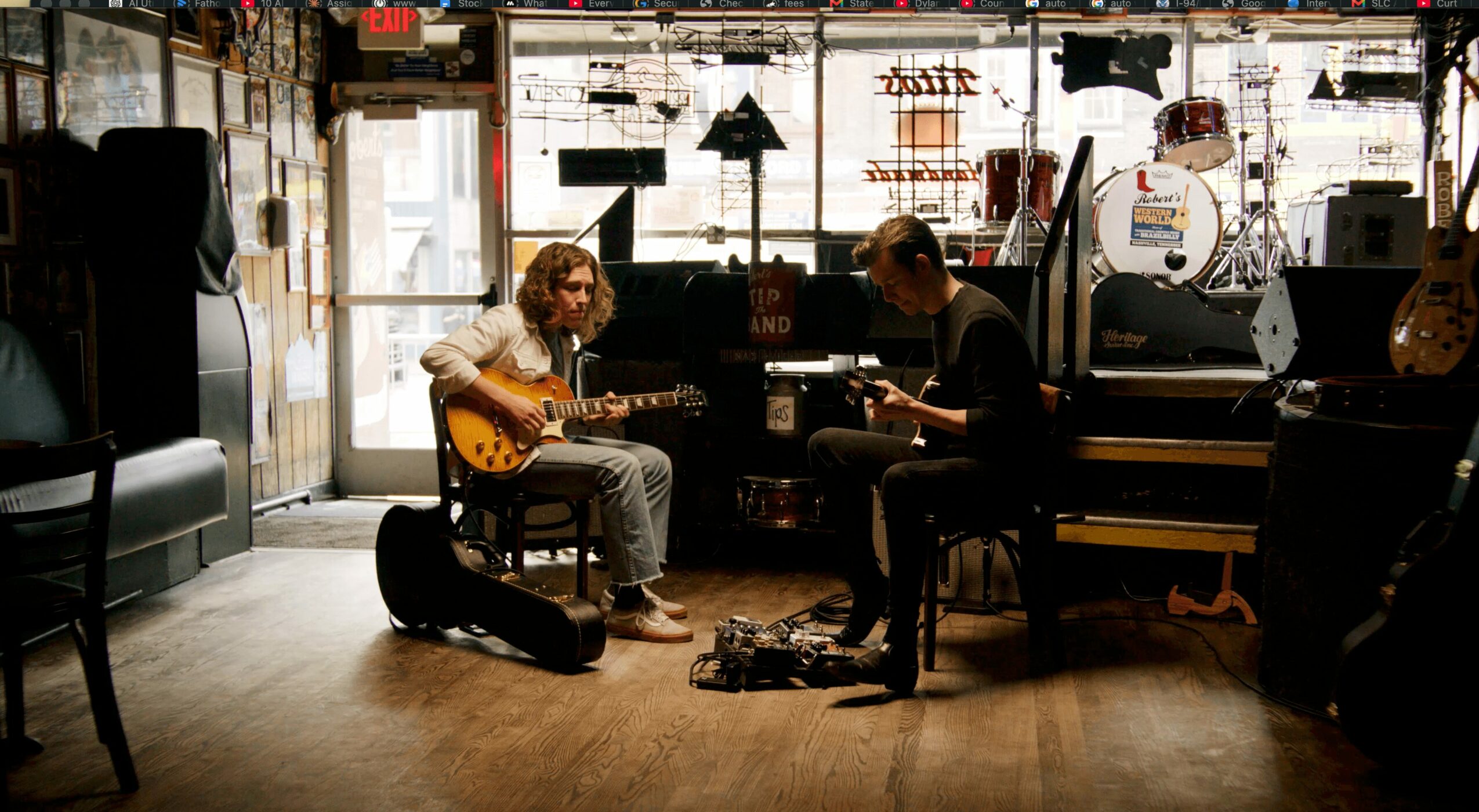 Two musicians playing guitars in a music studio with equipment and instruments around them.
